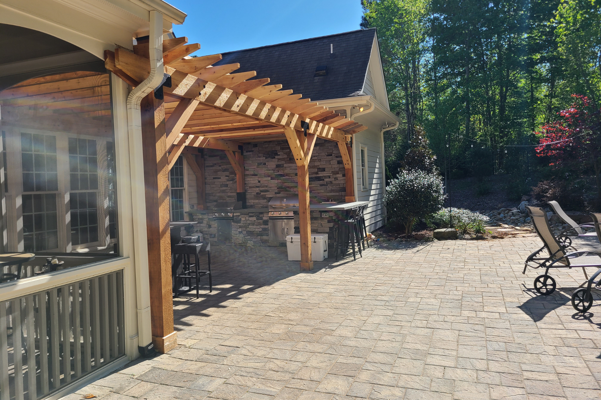 A stone patio features a wooden pergola covering an outdoor kitchen with a stone backsplash, grill, and bar seating.