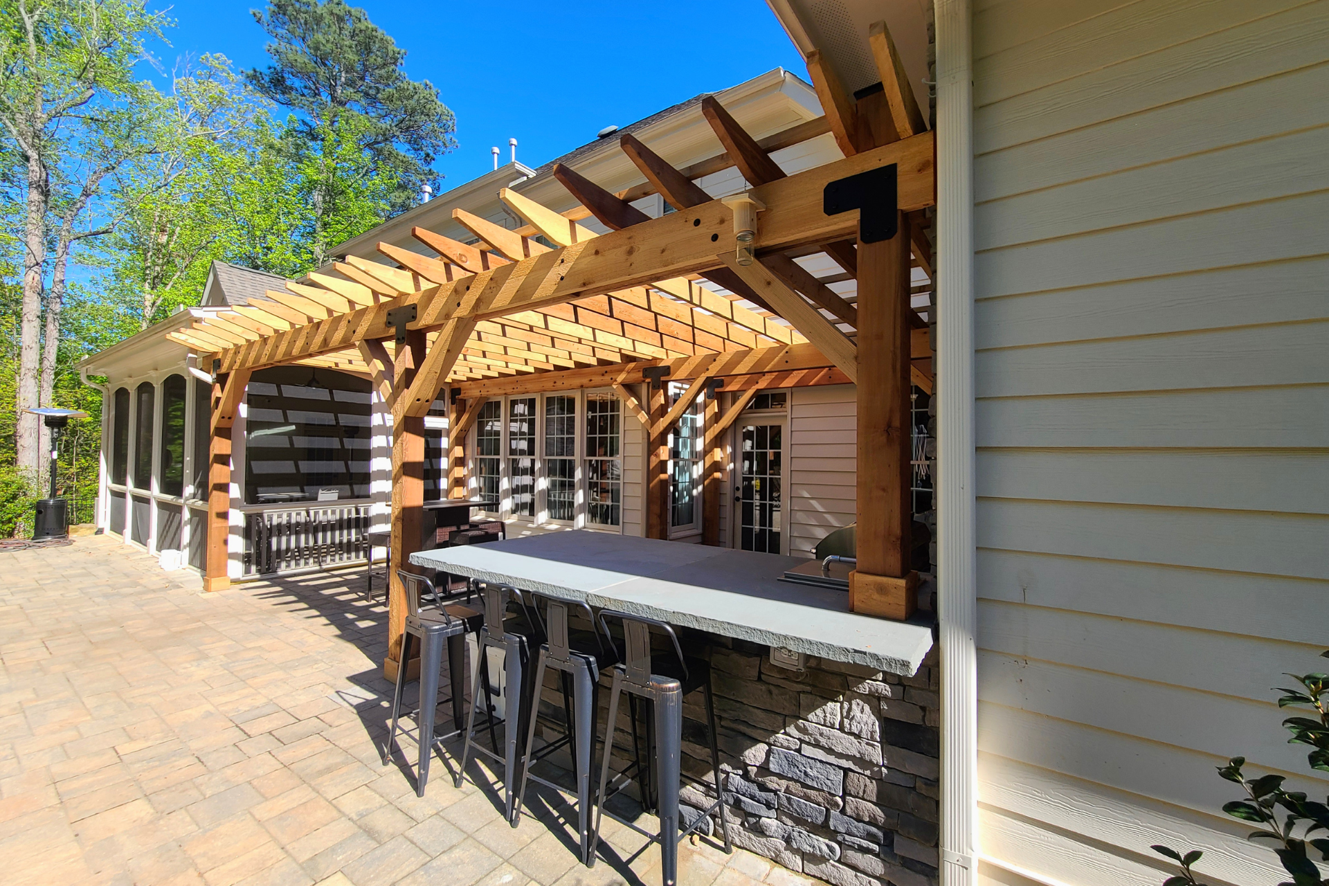 A wooden pergola covers a stone patio bar with four metal stools, situated next to a house with beige siding.