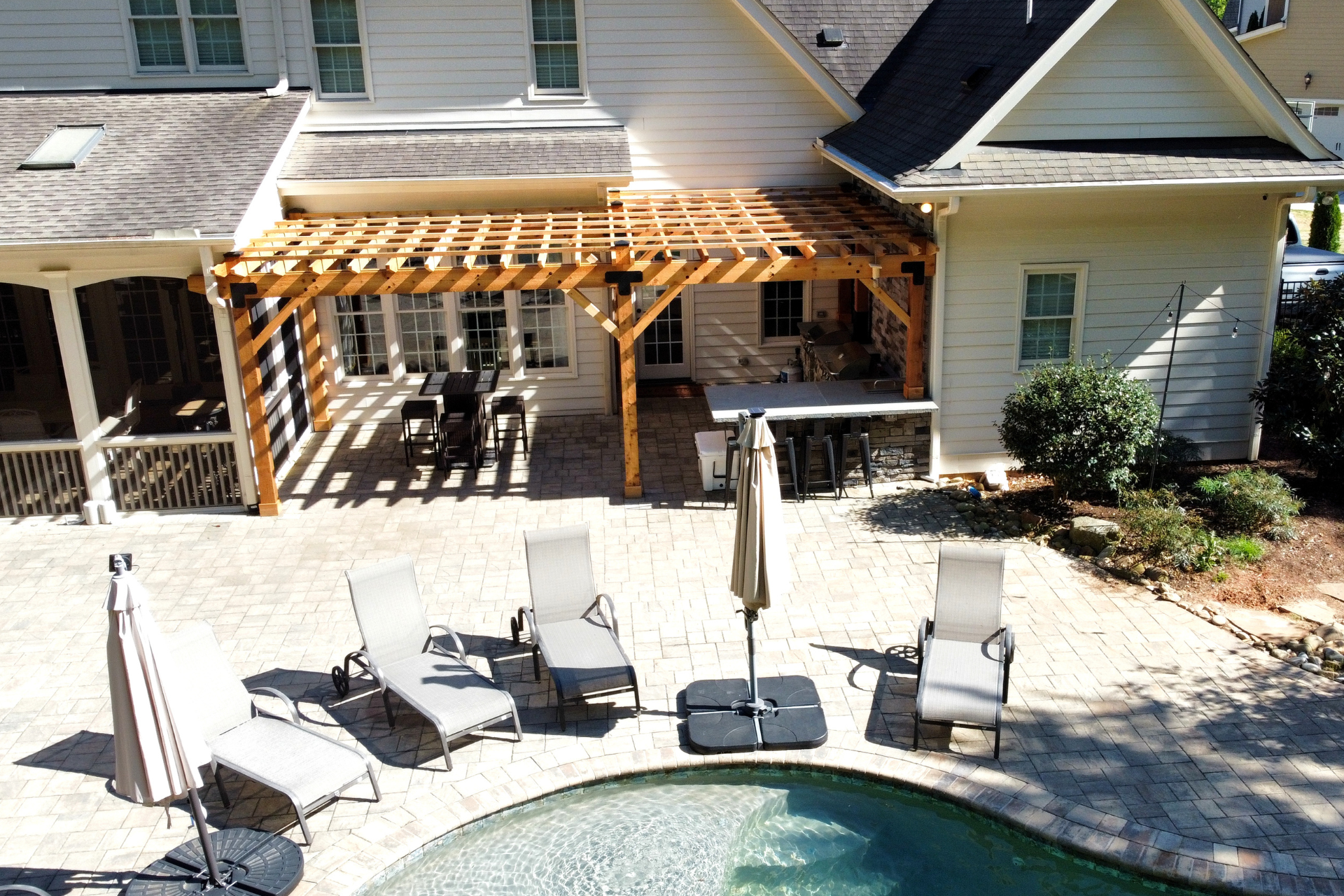 A patio with a wooden pergola, outdoor seating, and a pool, viewed from above next to a white house.