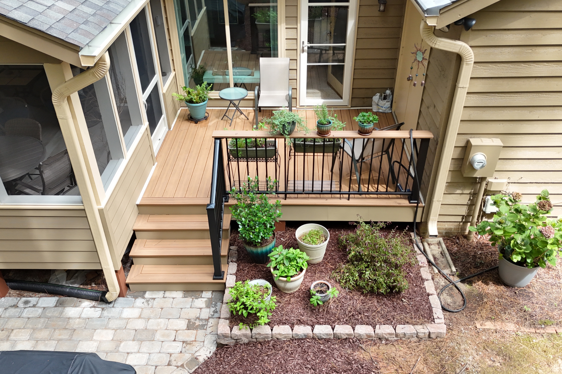 An elevated deck with stairs leads to a home exterior, featuring container plants and a garden bed in the mulched yard.