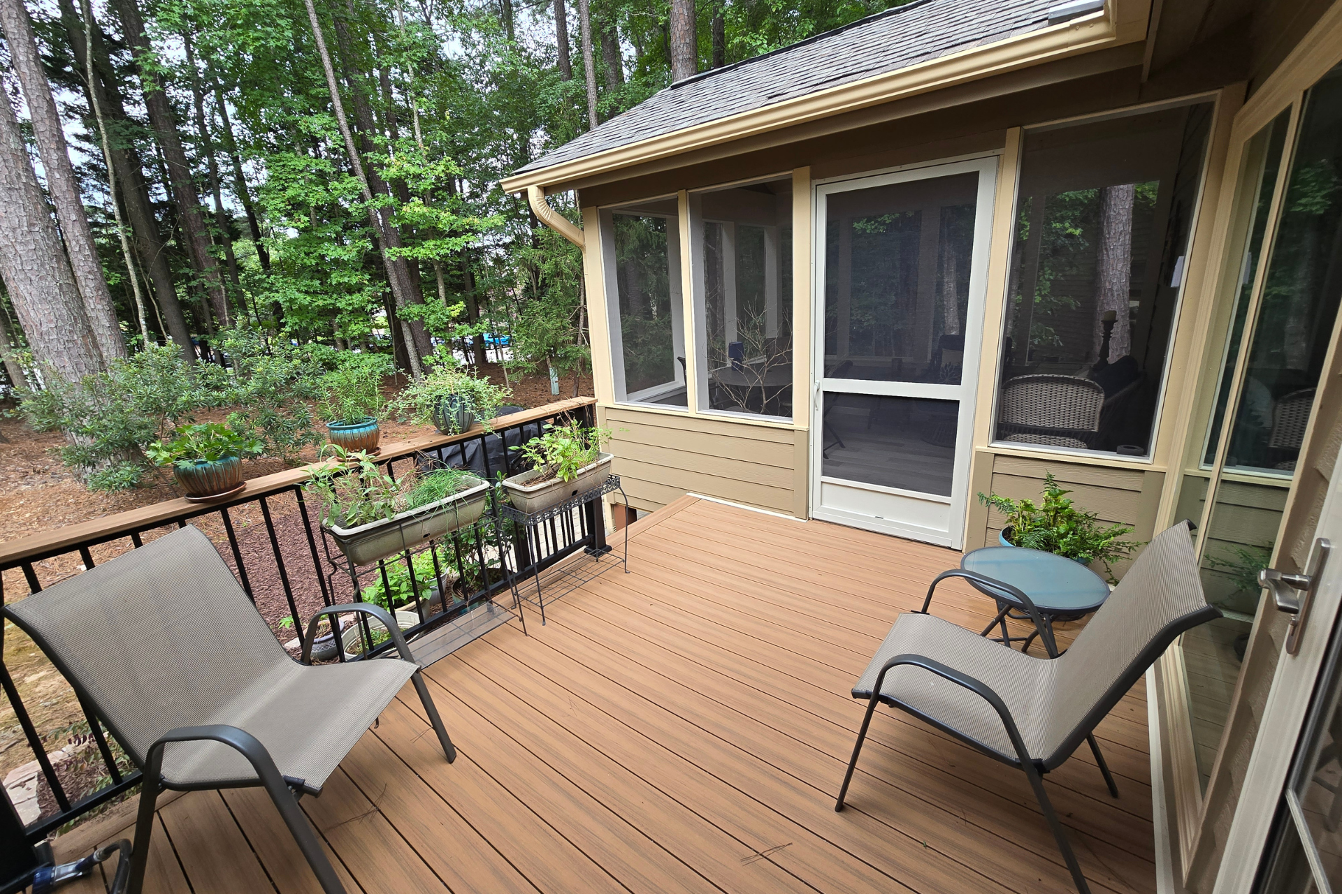 Two outdoor chairs and a small table sit on a wooden deck next to a screened-in porch with a view of a wooded area.