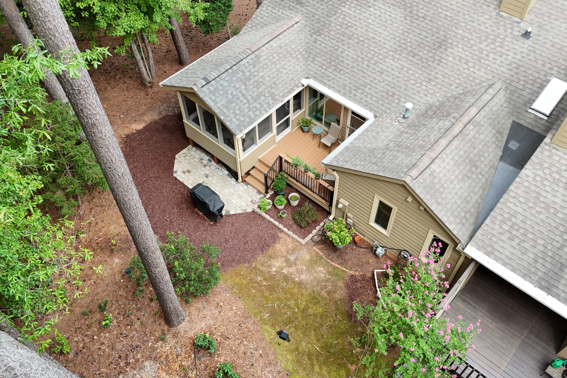 High-angle view of a beige house with a screened porch, wooden deck, and a yard with trees and mulch.