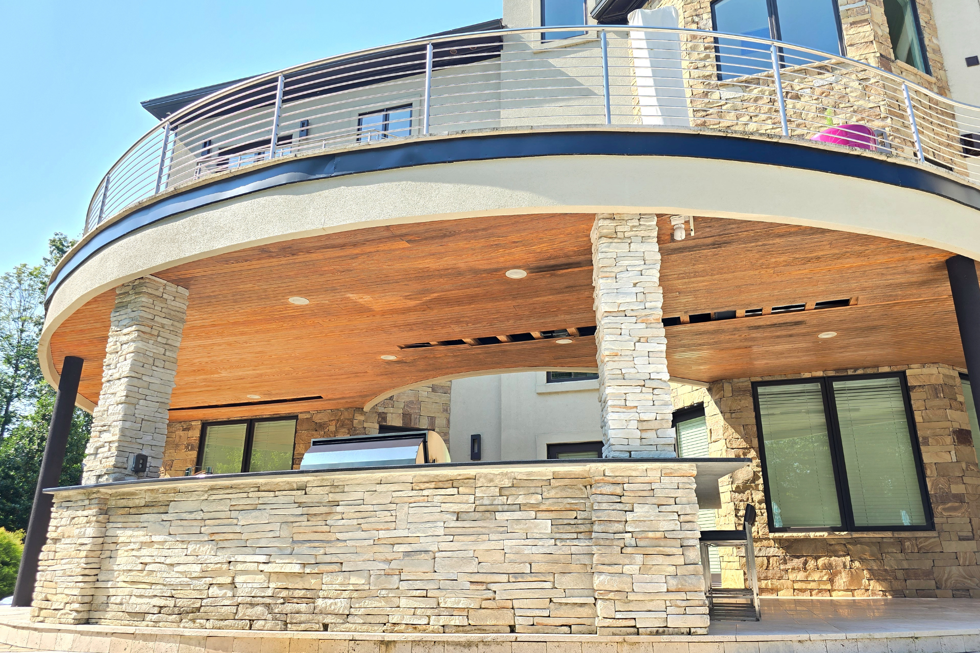 Outdoor stone kitchen island and patio beneath a large curved balcony with railings on a multi-story home.