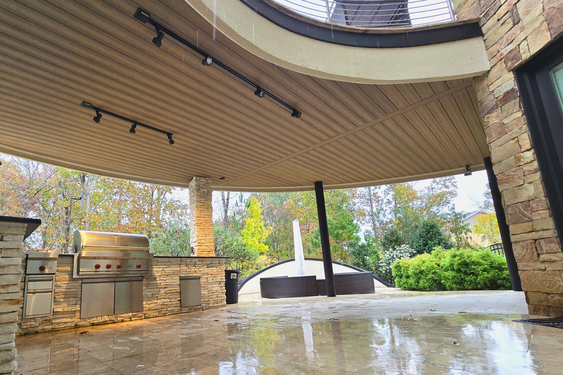 A covered stone outdoor kitchen with a stainless steel grill and a view of a landscaped backyard with a fountain.