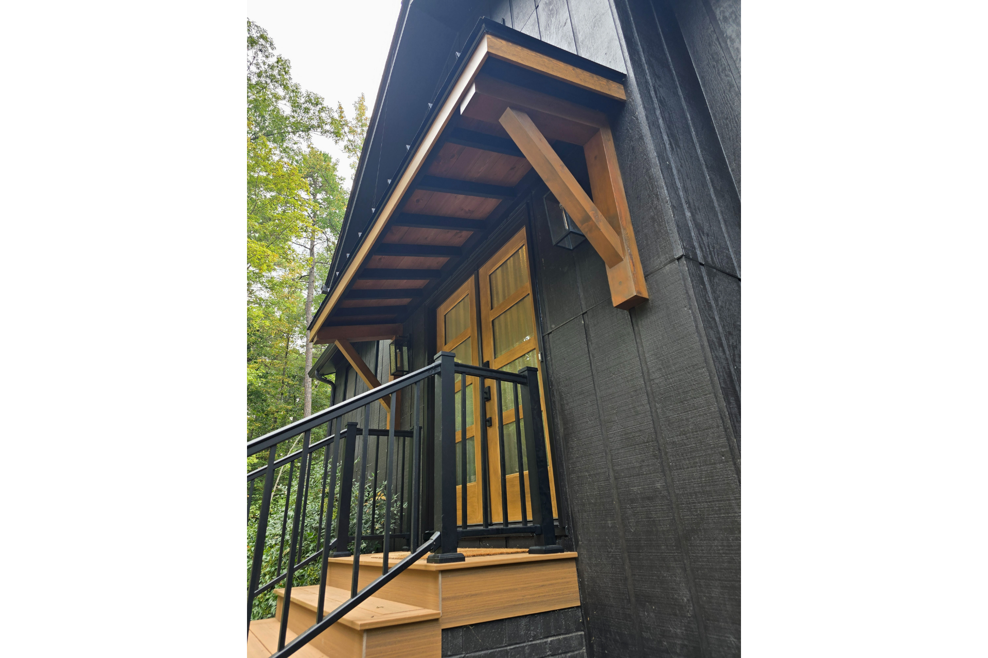 A low-angle view of a wooden entrance canopy above double glass doors, featuring black walls, railings, and deck stairs.