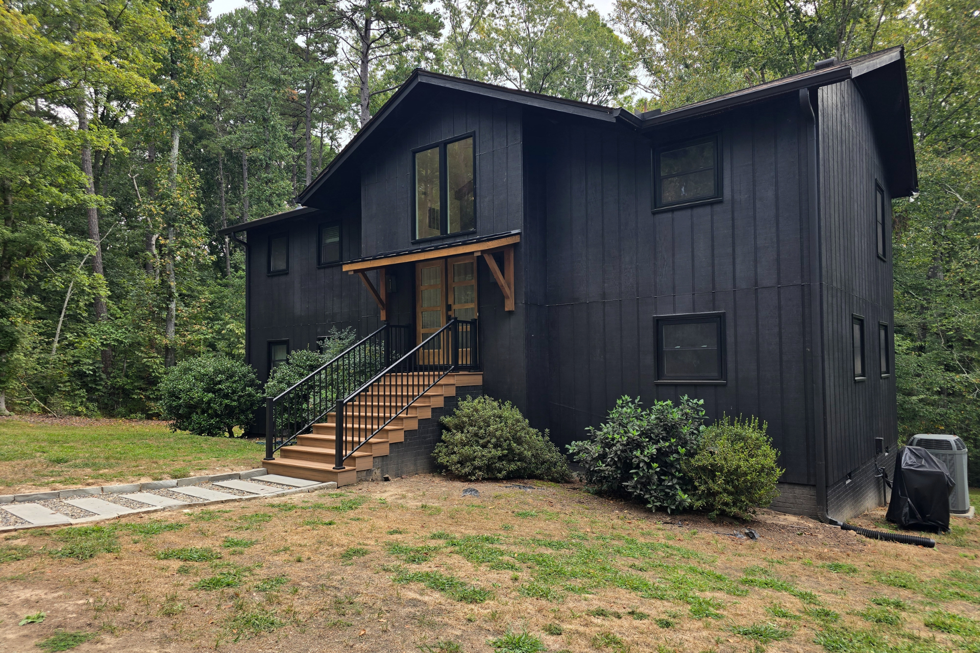 A two-story house with black board-and-batten siding and wooden front stairs, nestled among lush green forest trees.