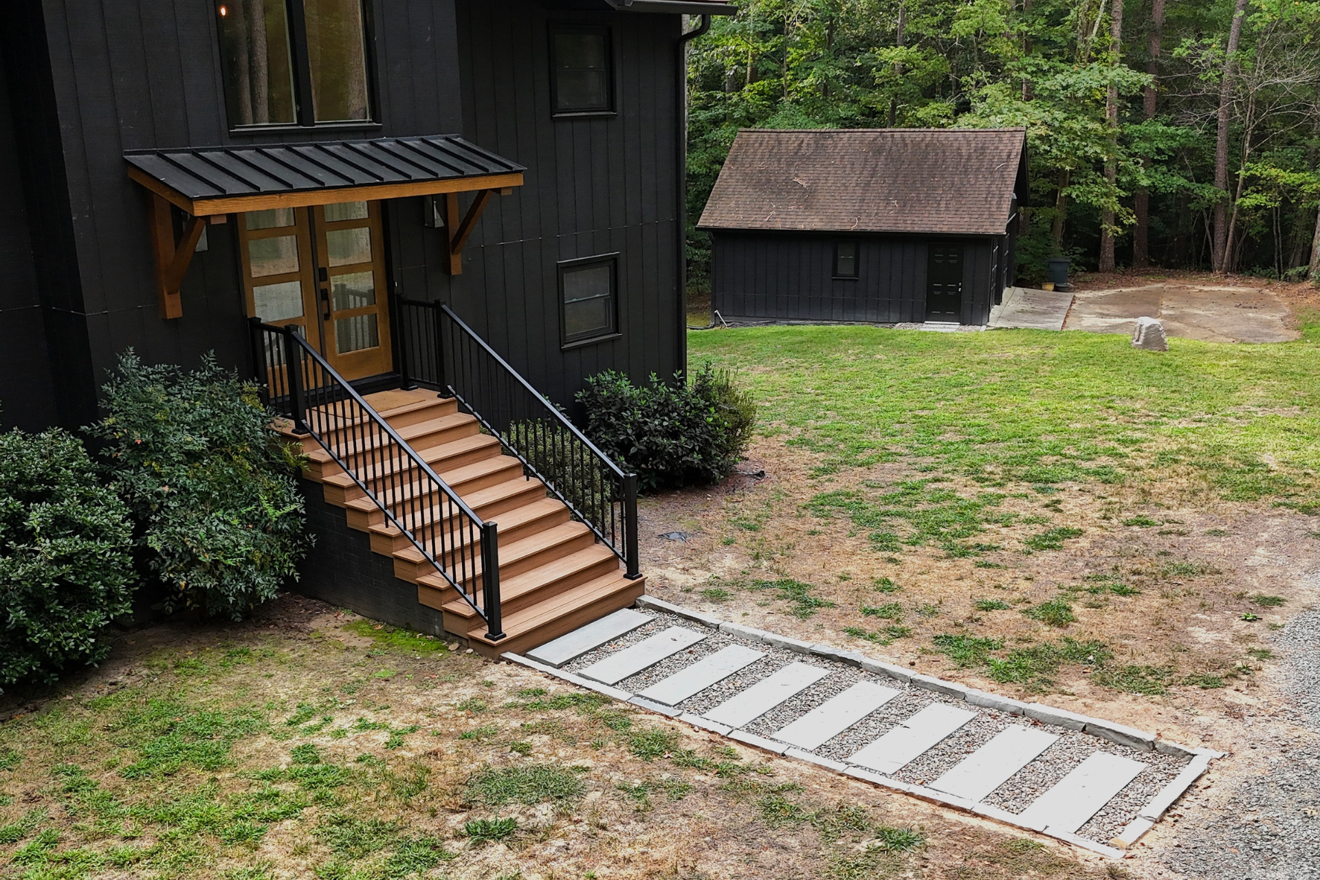 A black house with a wooden front staircase and a gravel path with concrete pavers leading to it in a wooded yard.