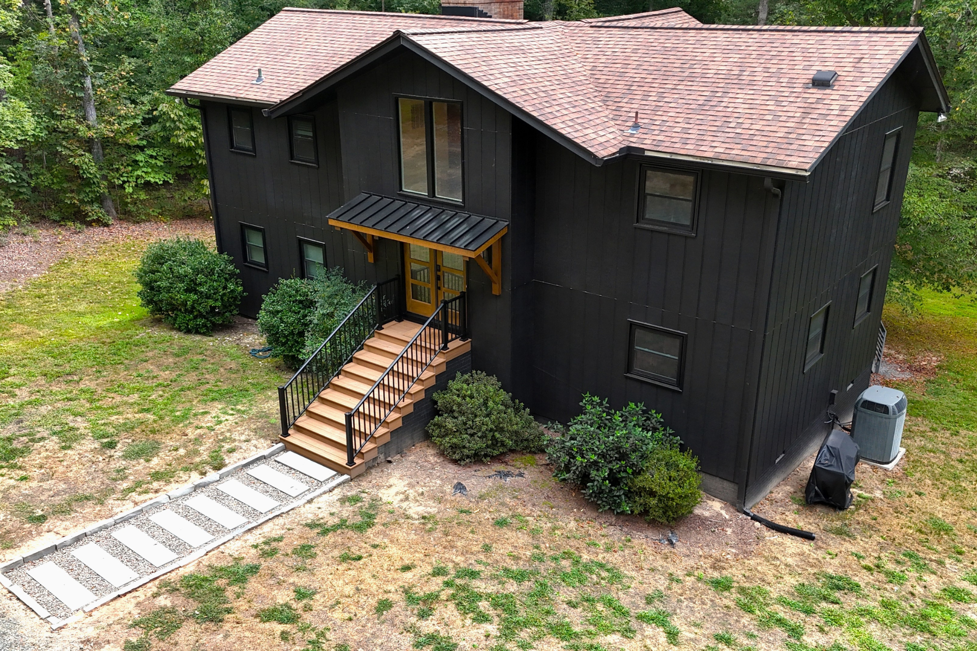 A two-story dark-sided house with a shingled roof, a front wooden staircase, and a stone walkway in a wooded yard.
