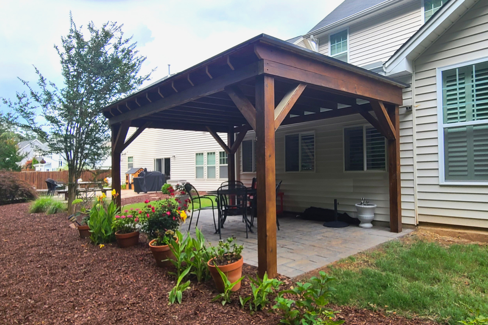 A wooden pergola stands on a paved patio beside a light-colored house, surrounded by a landscaped garden with mulch.