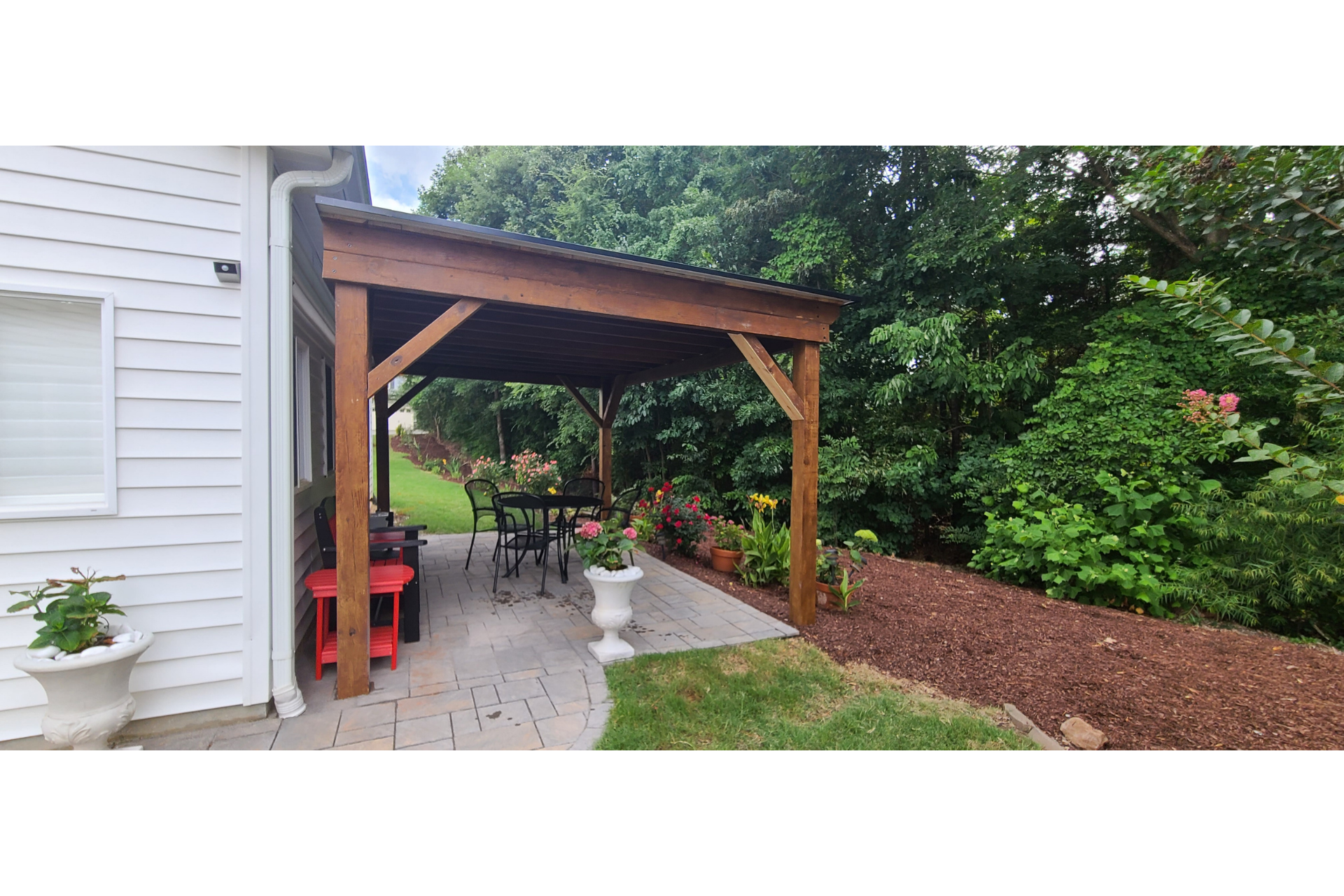 A wooden pergola covers a patio beside a white house, with a table, chairs, a red side table, and garden landscaping nearby.
