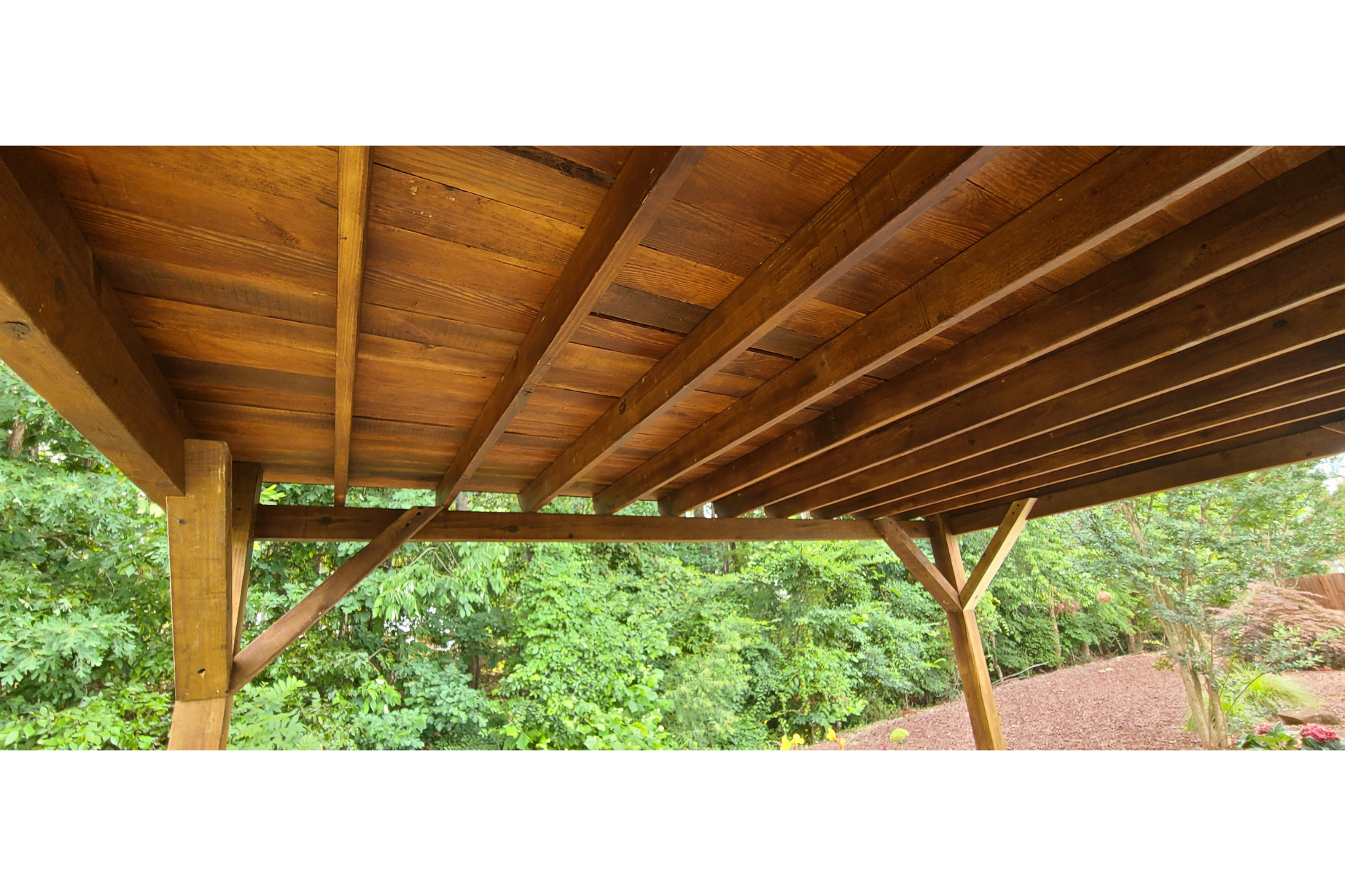 Underneath view of a brown wooden deck structure with joists, support beams, and diagonal braces, set against green trees.