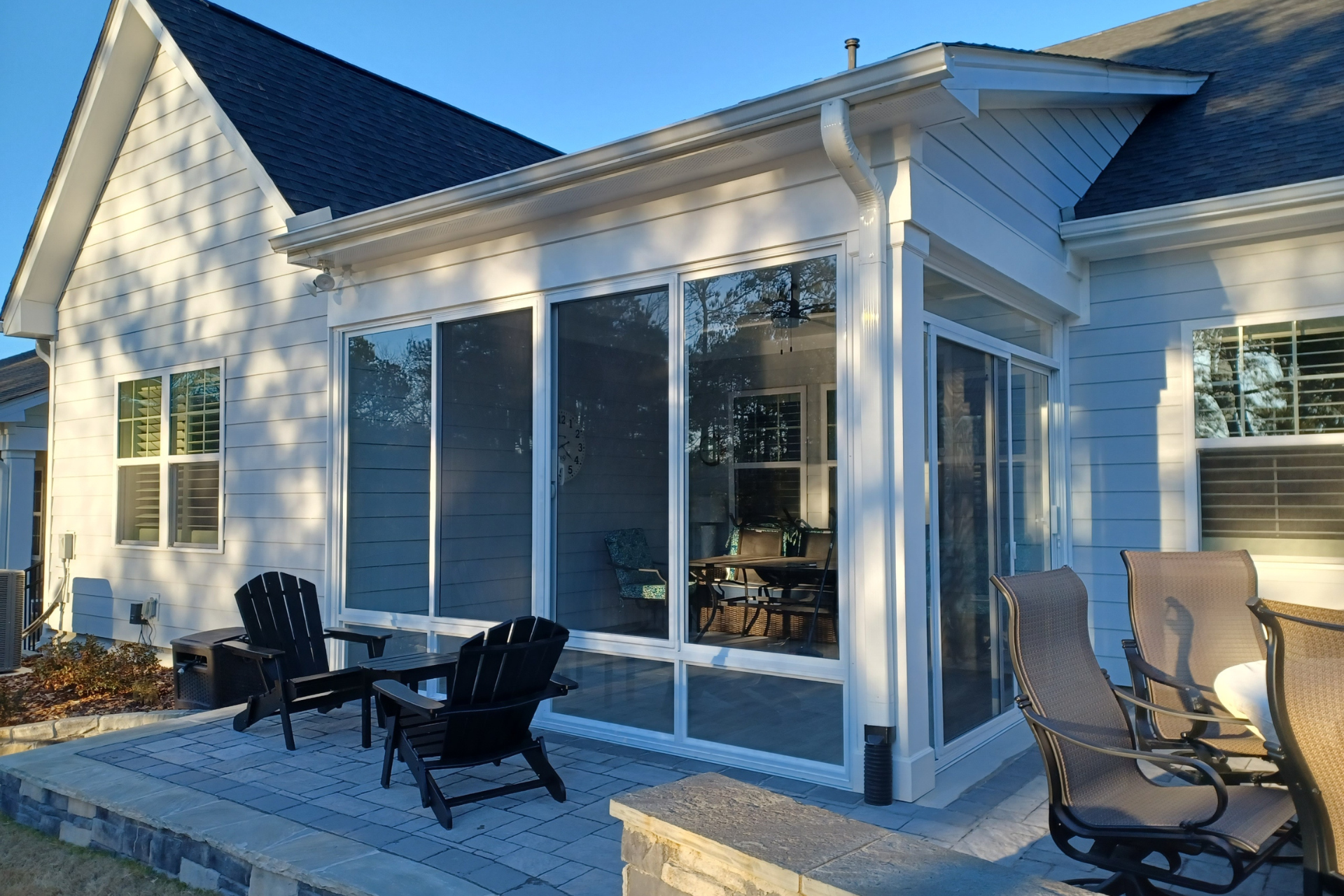 A white sunroom addition attached to a house, featuring large glass panels, a patio floor, and outdoor chairs.