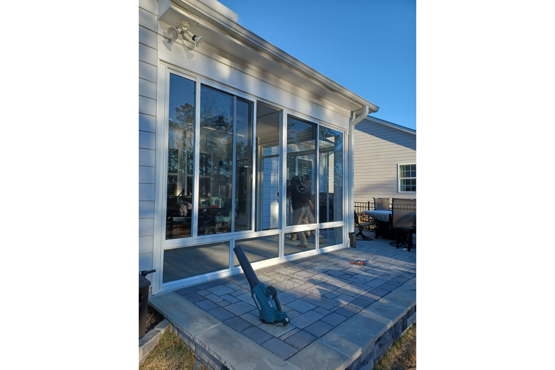 A leaf blower sits on a gray paver patio in front of a white sunroom with large glass windows and doors.