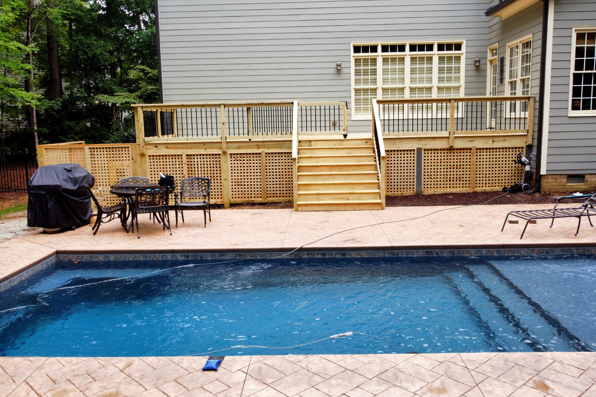 A rectangular backyard swimming pool with a patio and wooden deck, set against a two-story gray house with wood lattice.