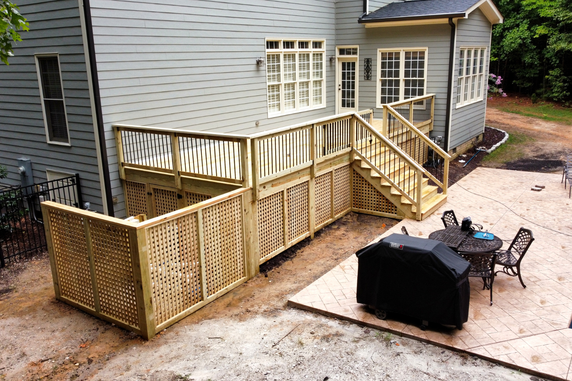 Newly constructed wooden deck with lattice skirting and stairs leading to a concrete patio with a grill and dining set.
