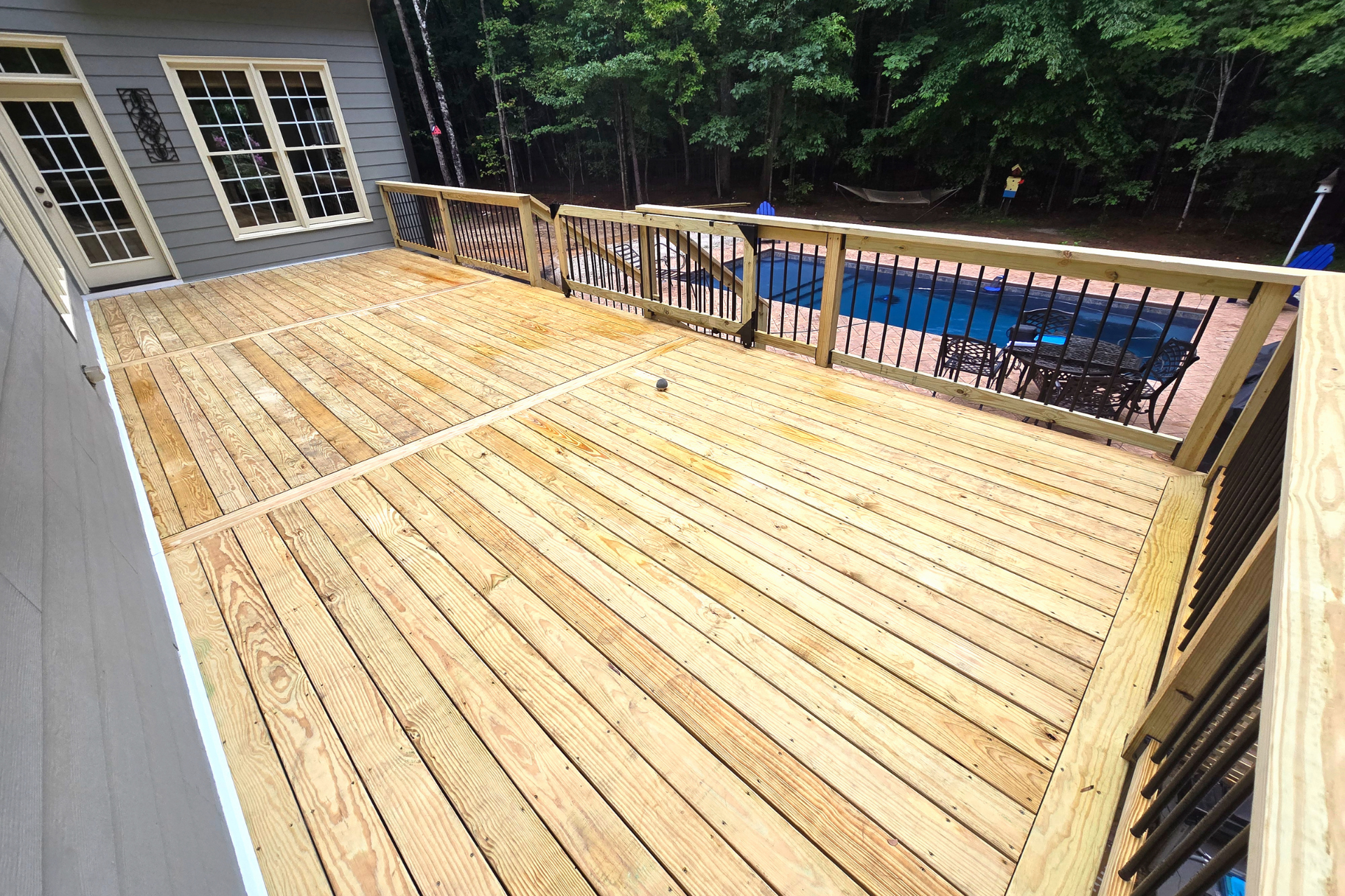 A high-angle view of a freshly stained wooden deck with black railings, overlooking a backyard with a blue pool cover.