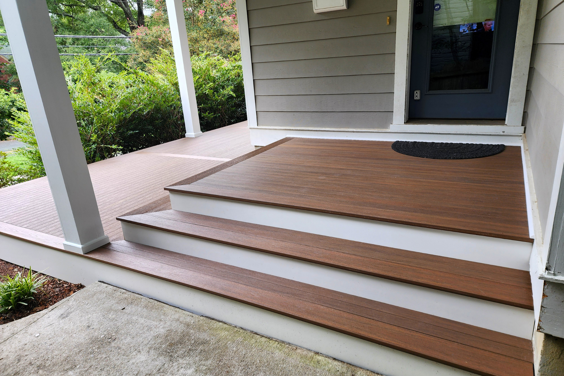 A front porch with wooden steps leading up to a dark-colored door, flanked by white support columns and a concrete walkway.