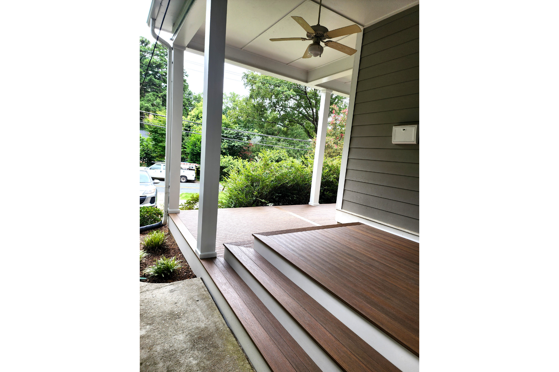 A covered porch featuring brown wooden decking, three steps, a ceiling fan, and grey siding, overlooking green trees.