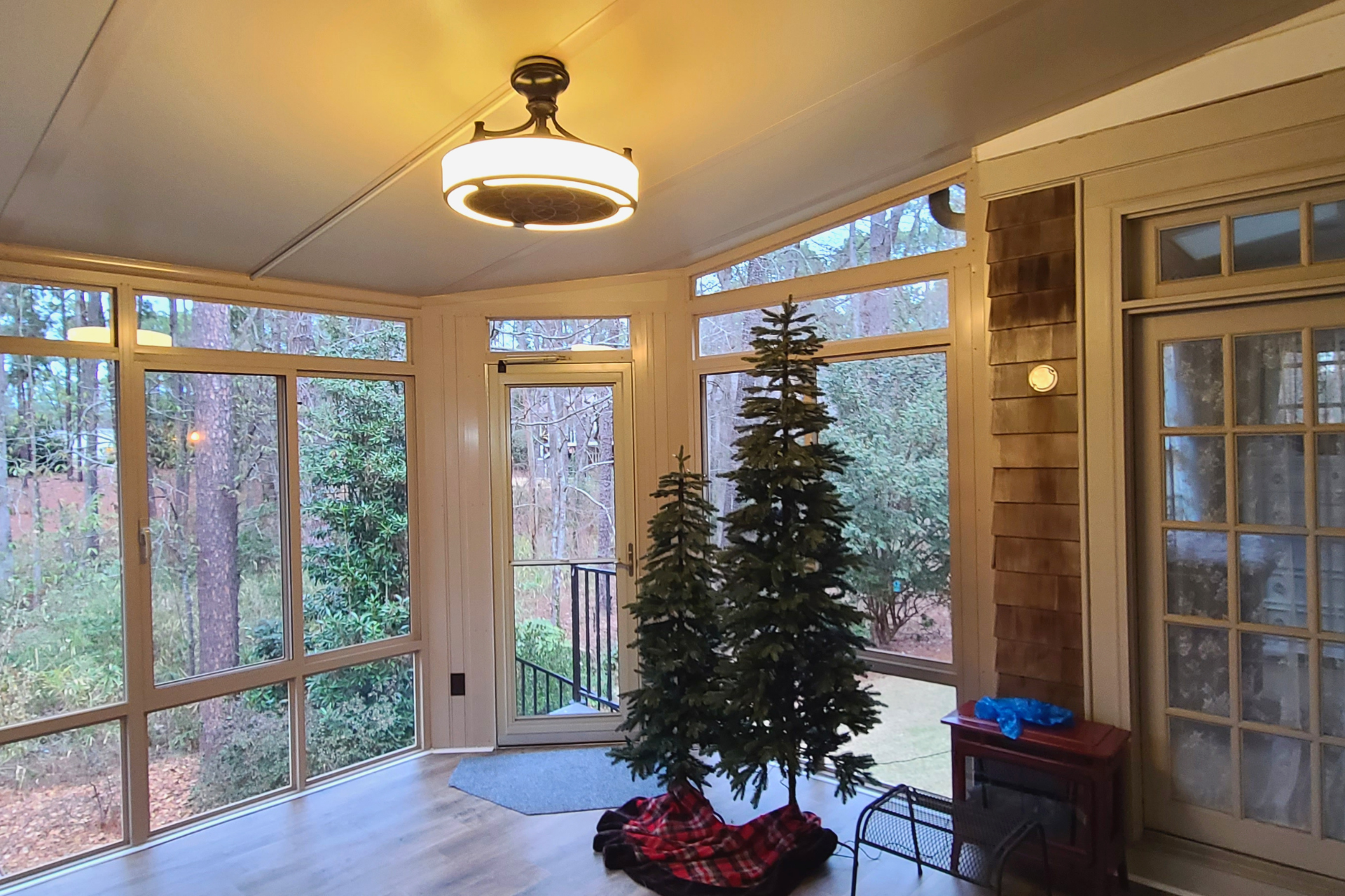 Sunroom with two unlit artificial Christmas trees, light wood floors, large windows, and a ceiling light fixture.