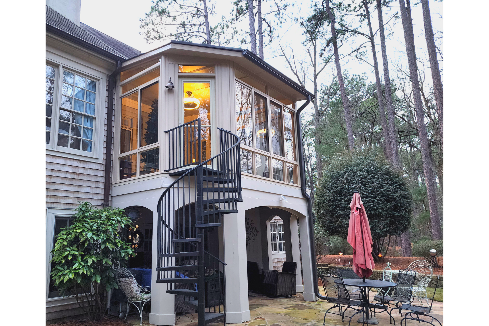 A two-story home extension with a sunroom above a patio, connected by a black metal spiral staircase in a wooded yard.