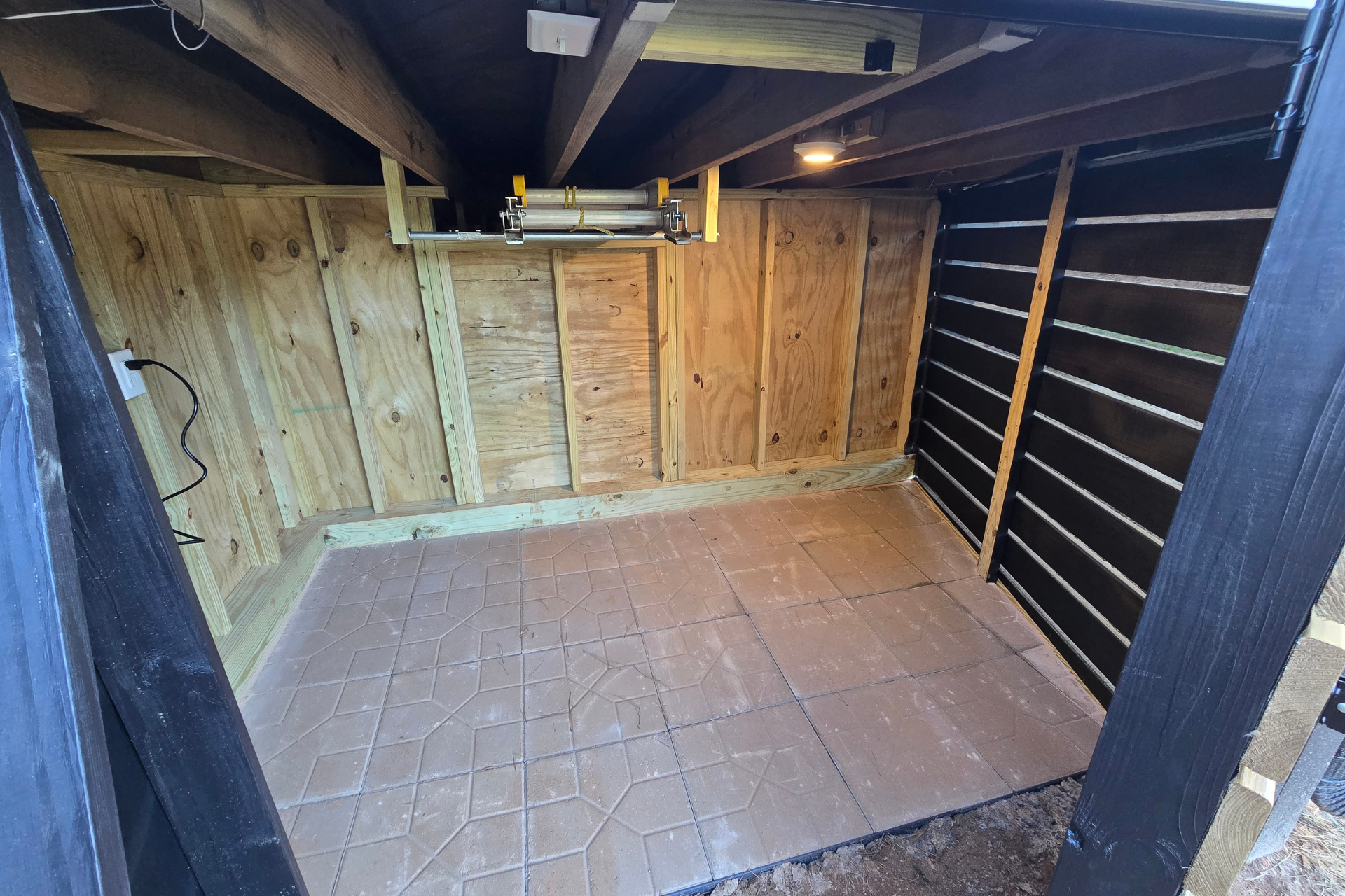 A small, open shed with plywood walls, a tiled floor, and exposed wooden ceiling beams.
