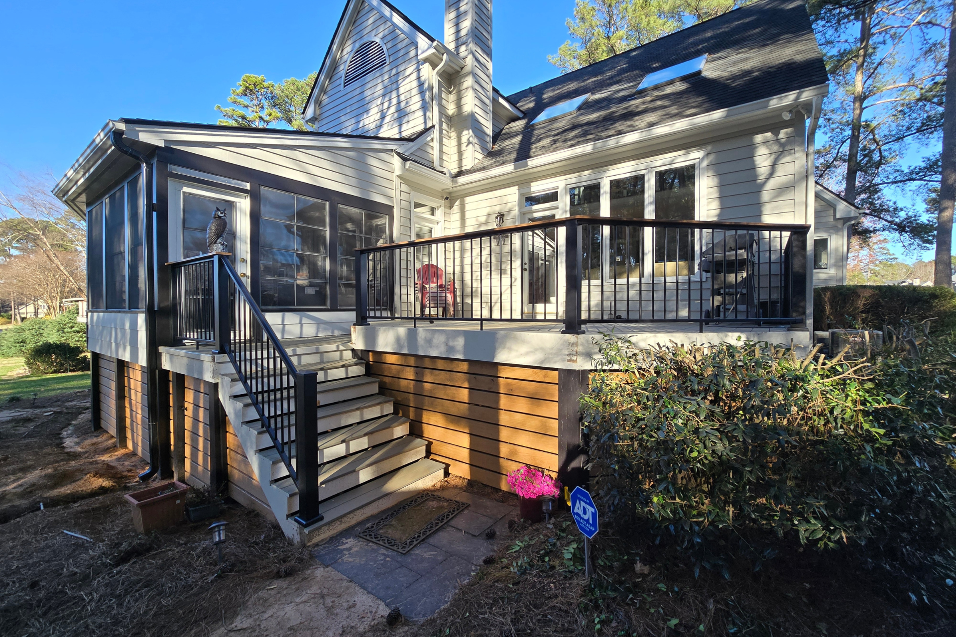 A white two-story house features a black-railed wooden deck, a screened-in porch, and outdoor stairs over a lawn.