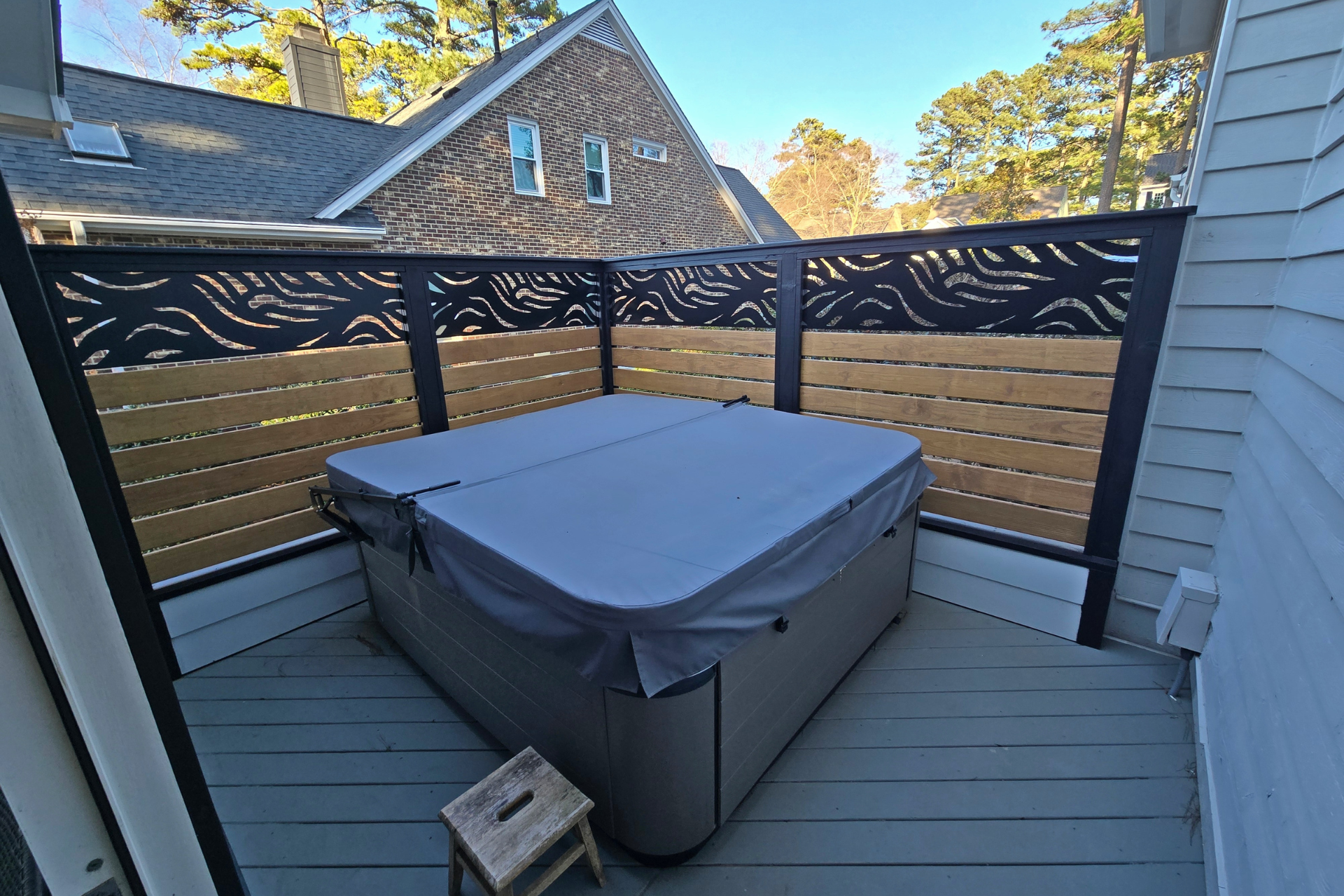 A grey hot tub with a cover sits on a deck, enclosed by a wooden privacy fence with black decorative metal panels.