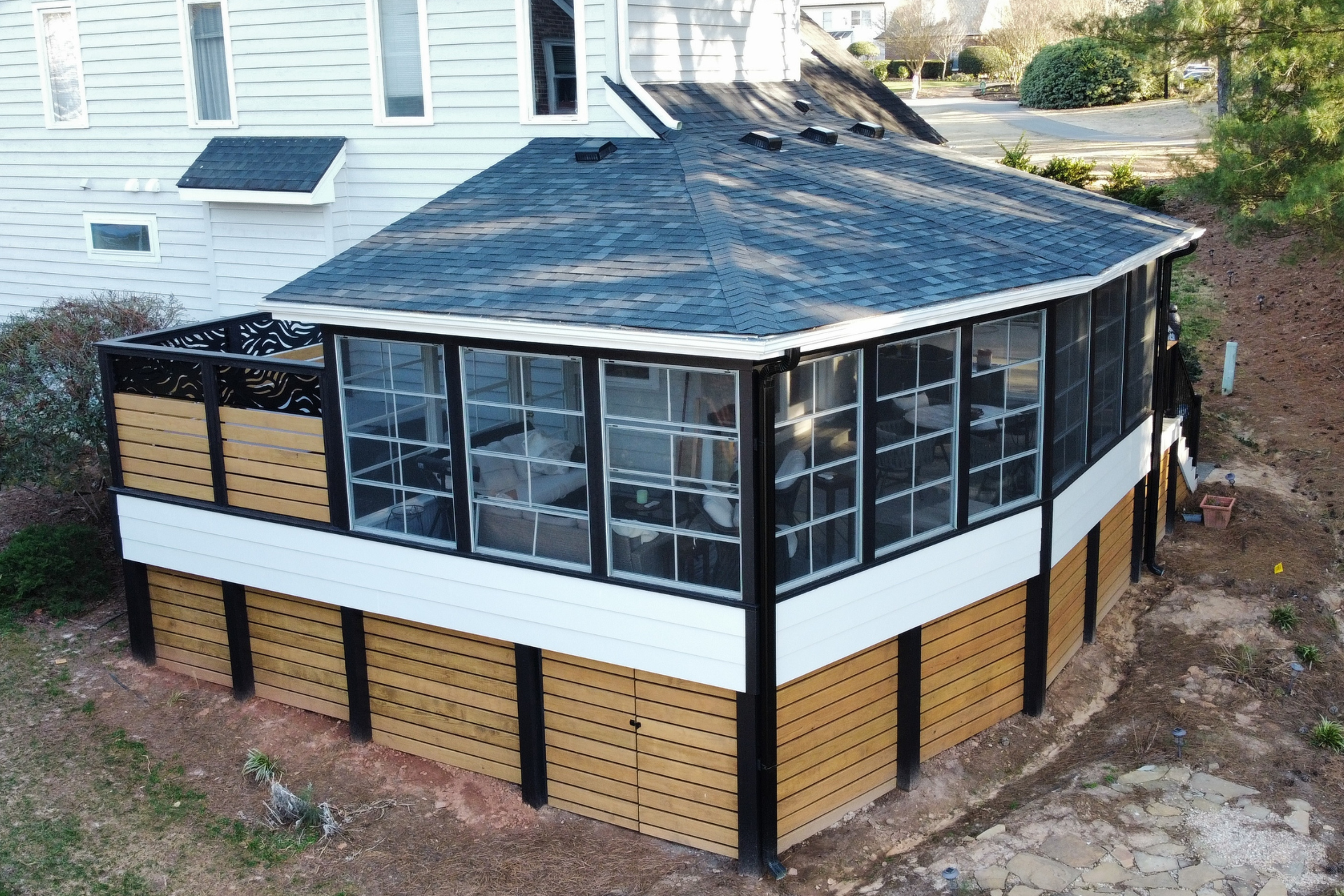 An elevated sunroom addition with a dark shingled roof, black framing, and horizontal wood skirting attached to a house.