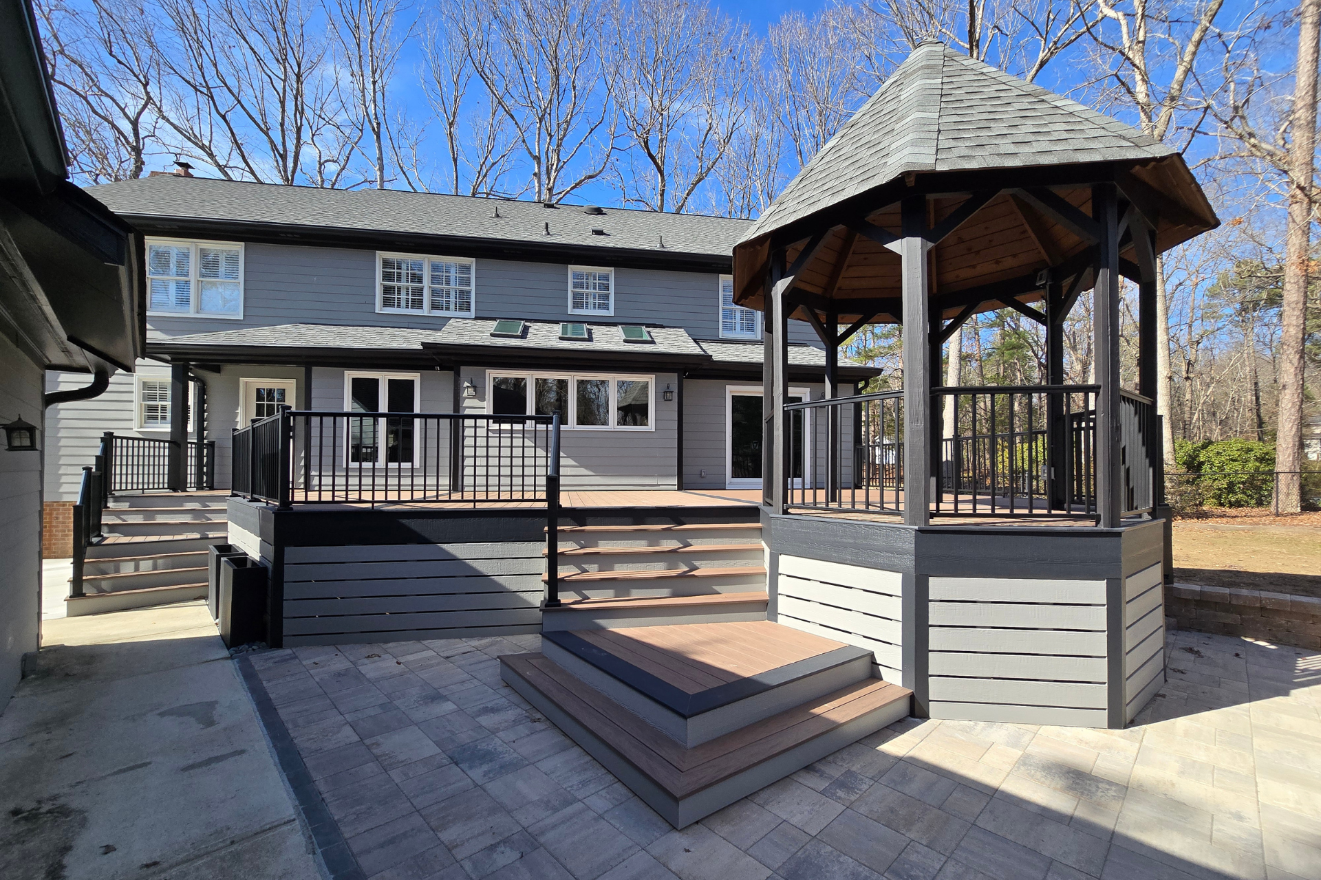 A grey two-story house features a large composite deck, stairs, and a matching octagonal gazebo in a wooded backyard.