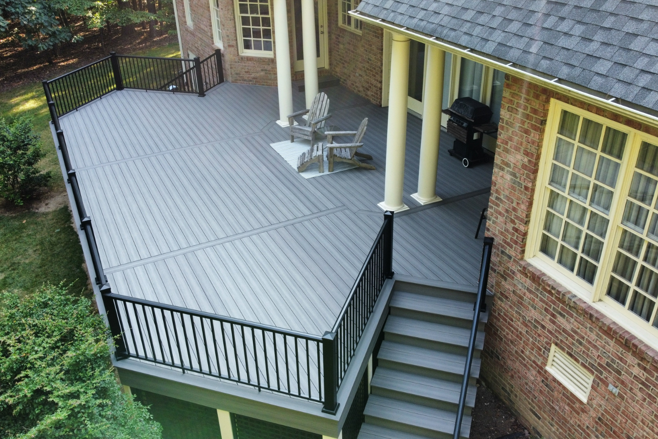 An elevated gray composite deck with black railings and stairs, attached to a brick house with white columns.