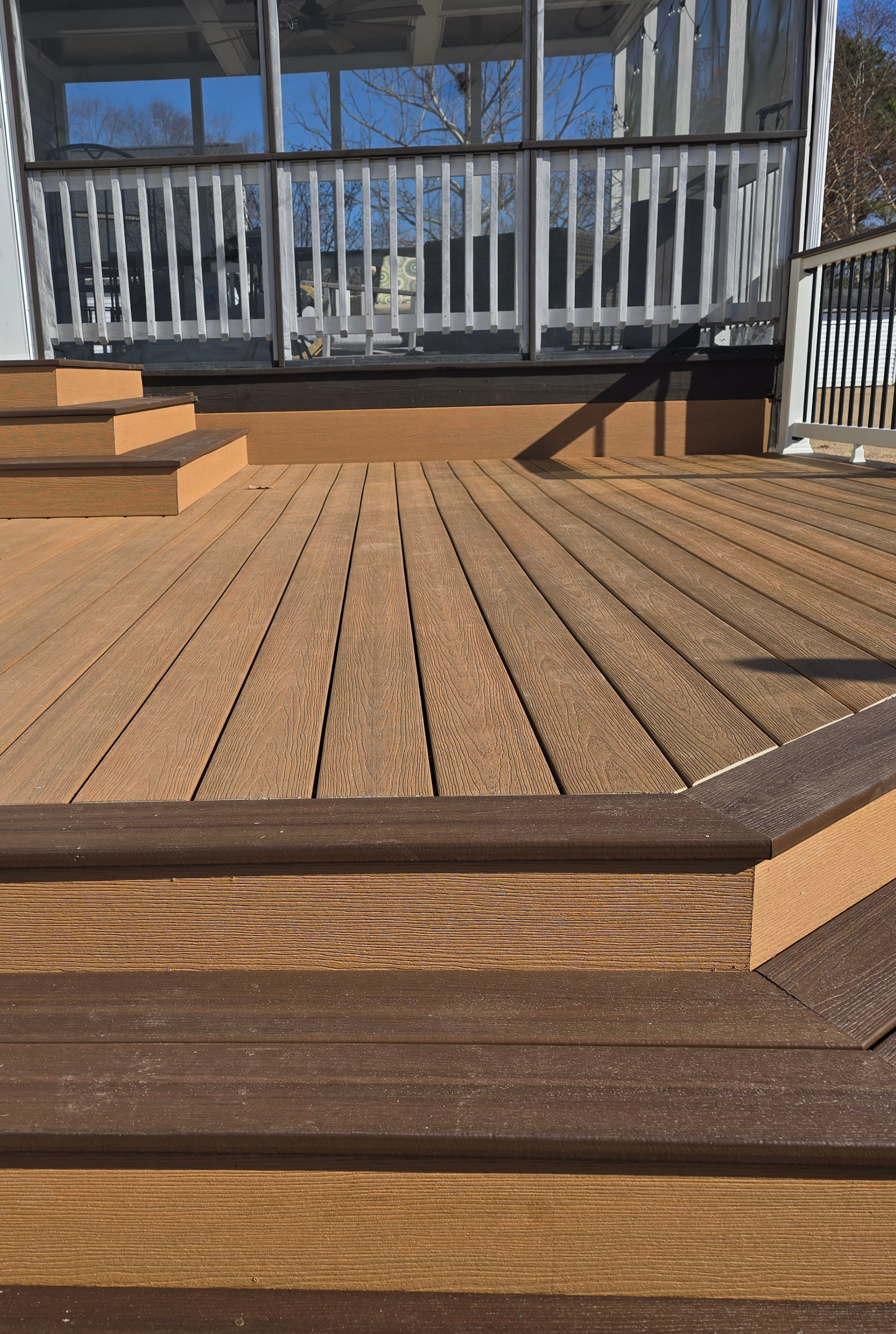 A multi-level outdoor deck with light tan composite decking, dark brown trim, and steps leading to a screened-in porch.