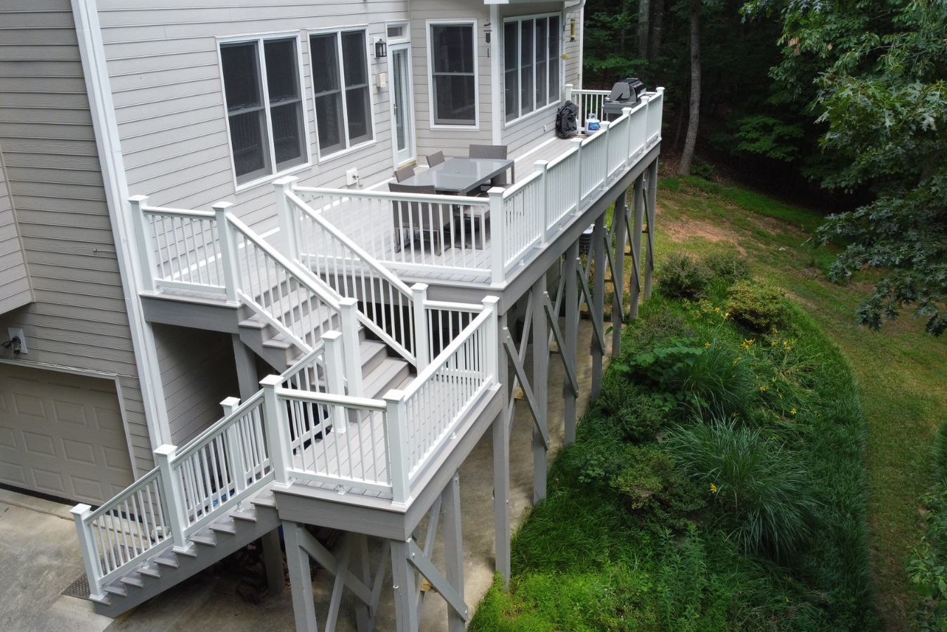 An elevated gray deck with white railings and stairs leads to a house exterior, overlooking a lush, sloped backyard.
