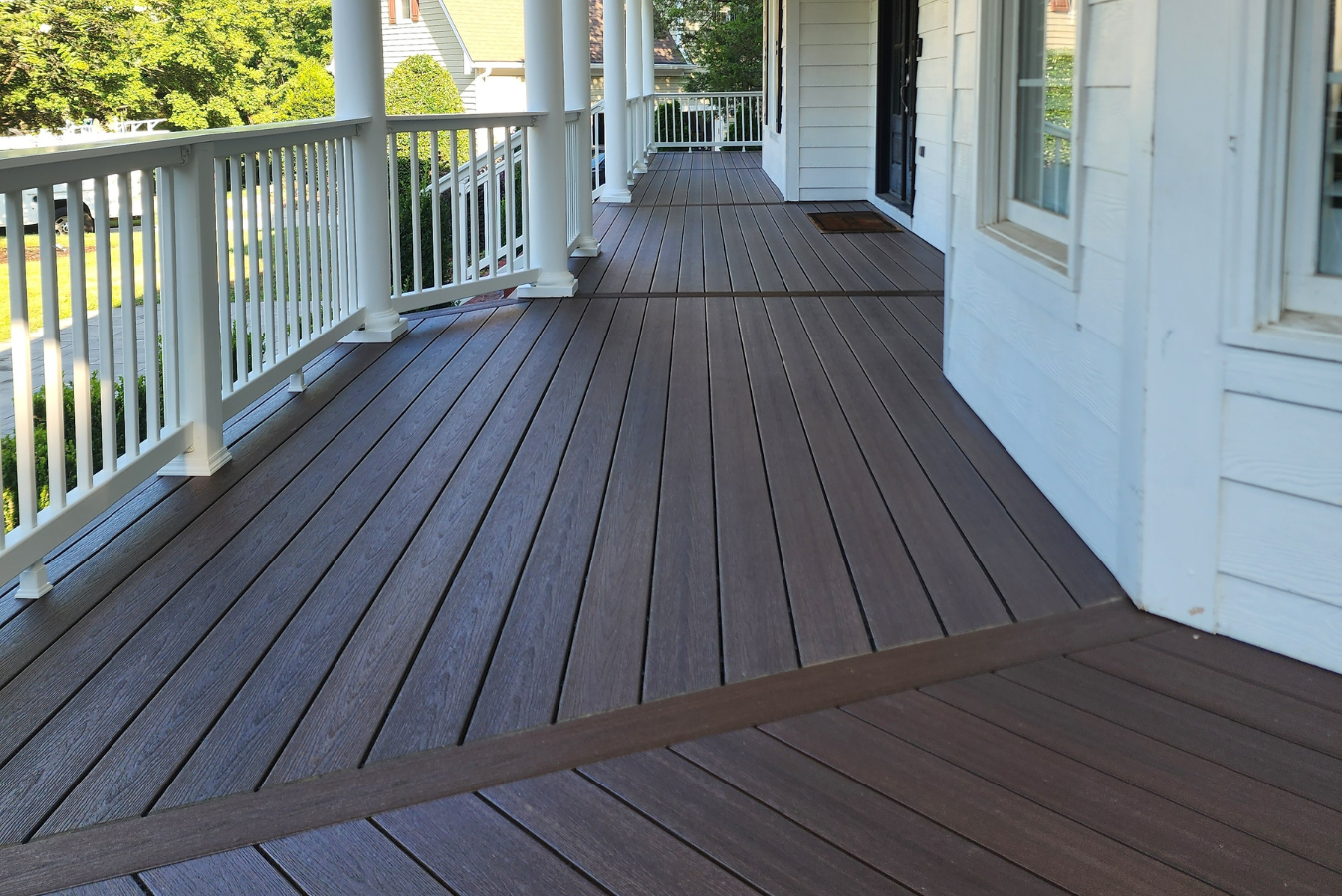 A wooden deck porch with white railings and columns extends along the side of a white house.