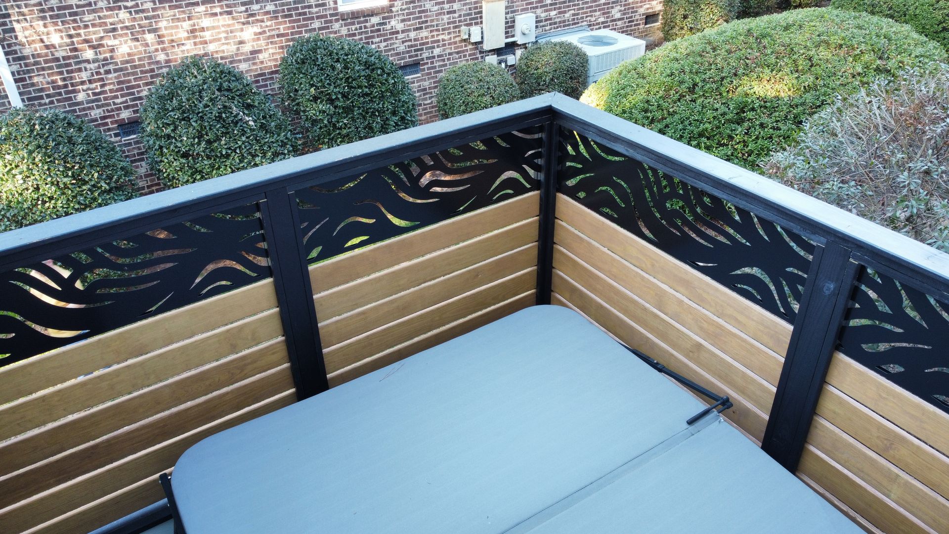 A hot tub covered by a grey lid is enclosed by a privacy fence with wood-look horizontal slats and black patterned panels.