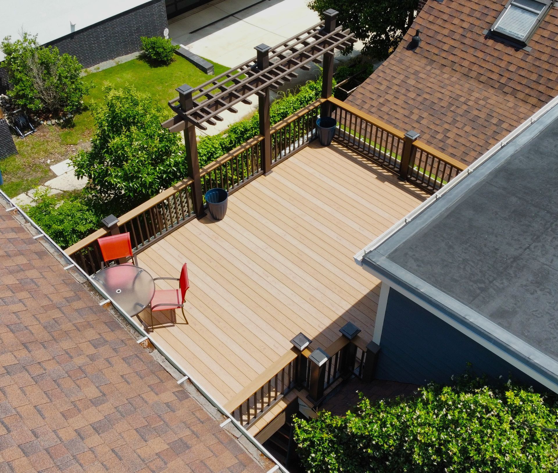 An aerial view of a wooden deck with a pergola, railings, two chairs, and a table, nestled between two house roofs.