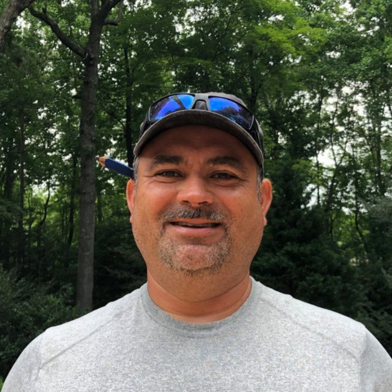 A person wearing a grey shirt and a baseball cap with blue lenses, smiling outdoors against a background of trees.