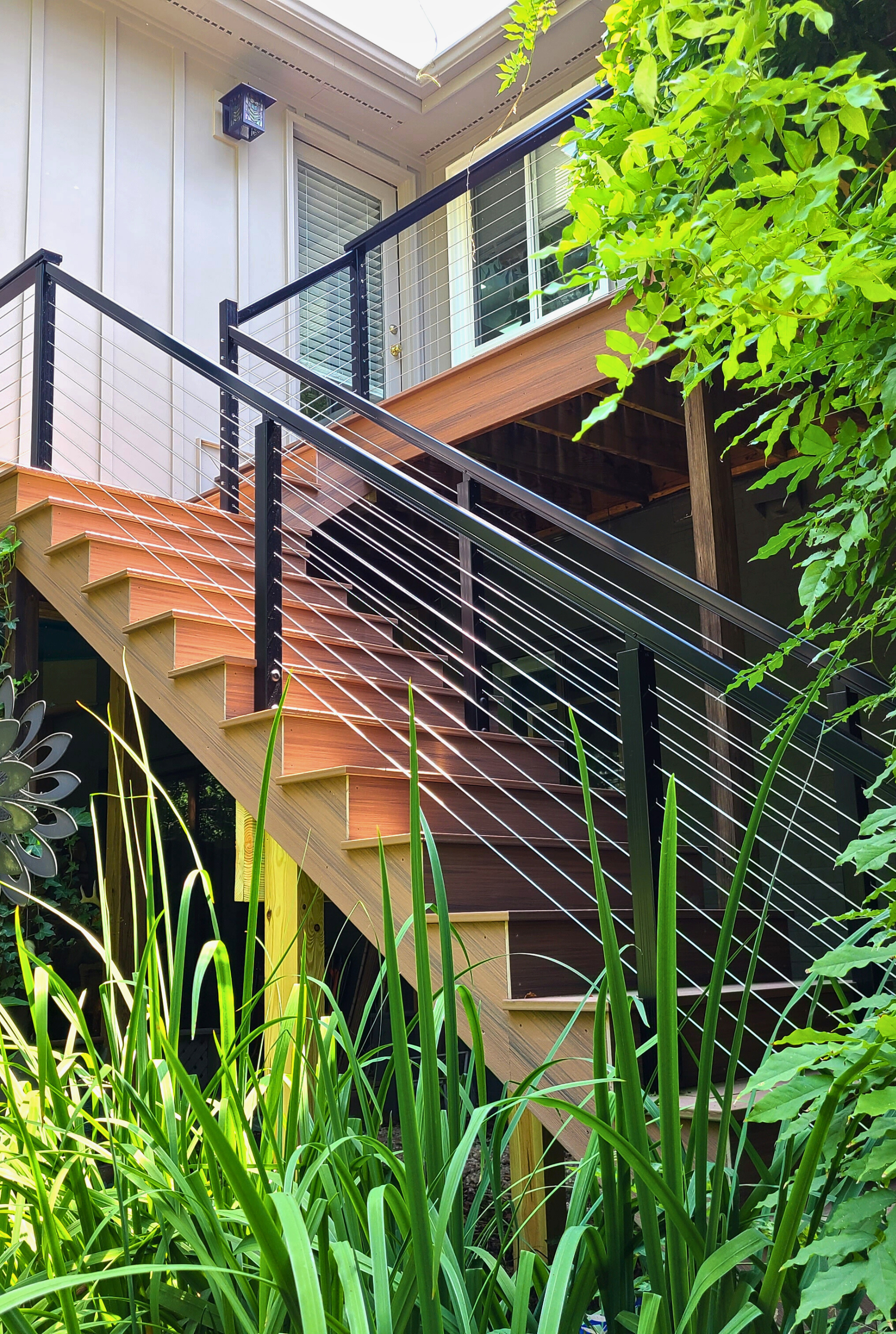 Wooden outdoor staircase with black metal railing and cable infill, rising toward a house deck surrounded by greenery.