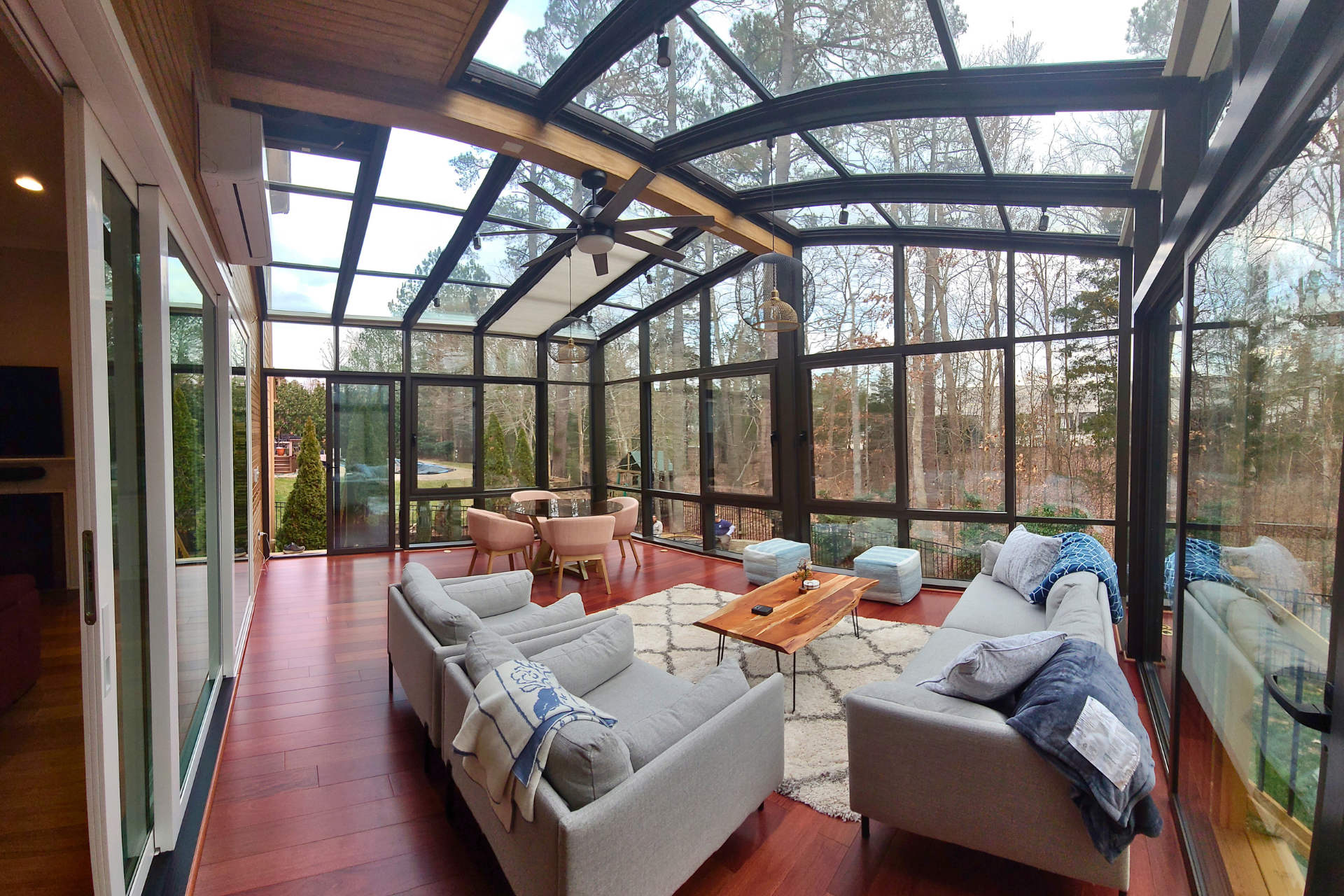 A sunroom with a glass roof, wooden floors, two light grey sofas, a small wooden coffee table, and an outdoor view.