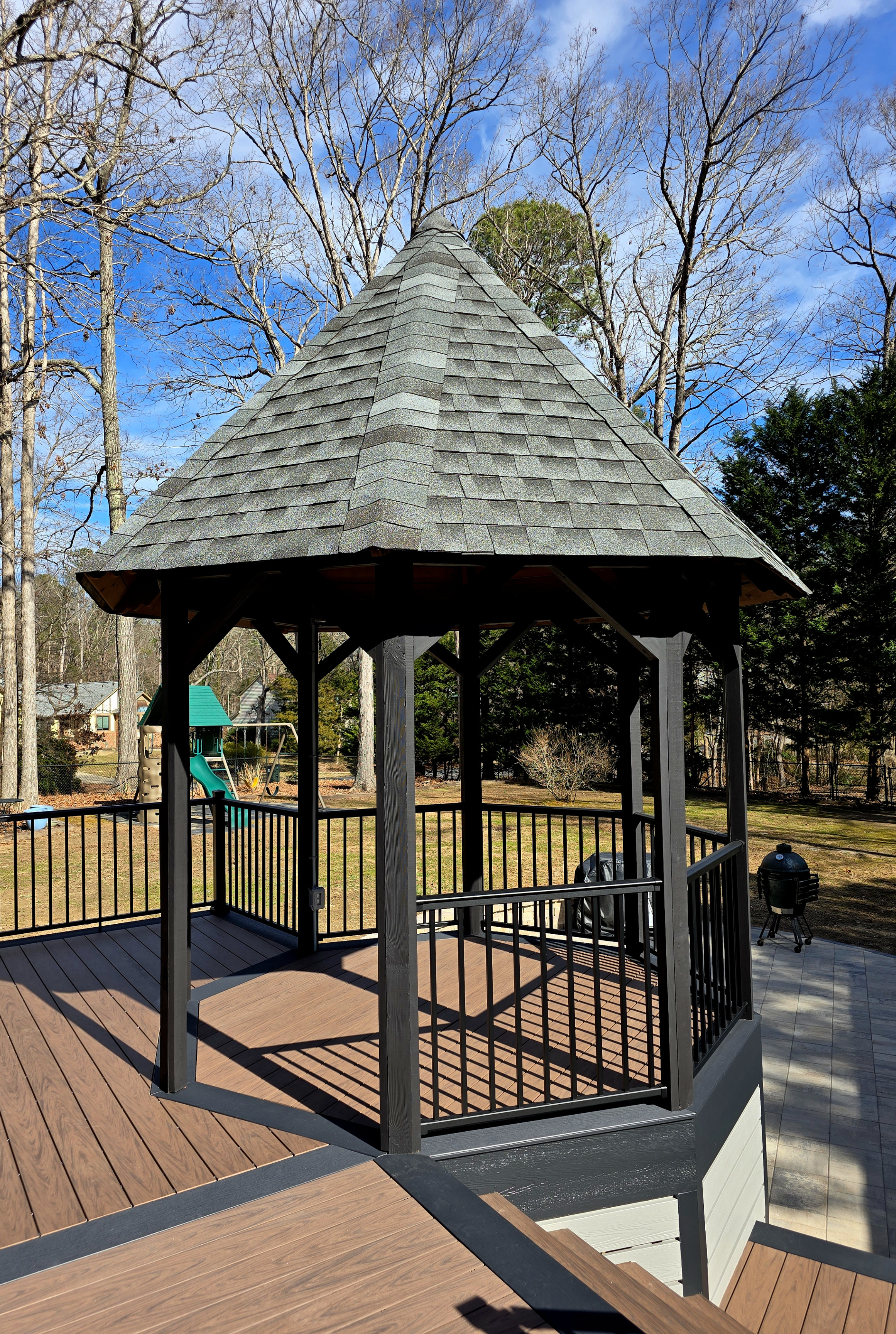 A dark-framed wooden gazebo with a gray shingled roof stands on a composite deck in a wooded backyard setting.