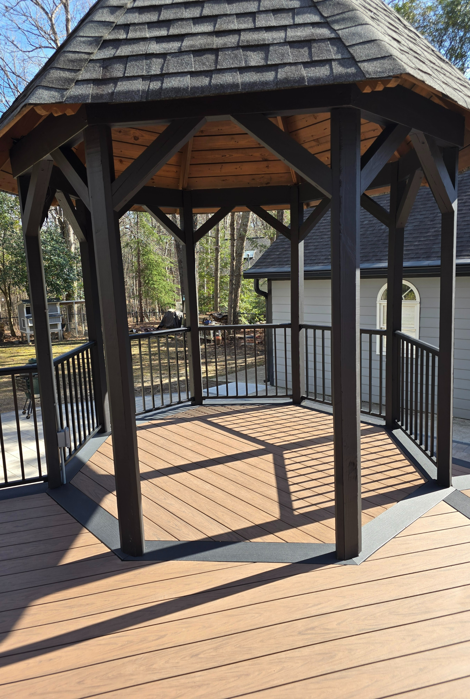 A wooden gazebo with dark-stained posts and a shingled roof stands on a composite deck in a sunny outdoor setting.