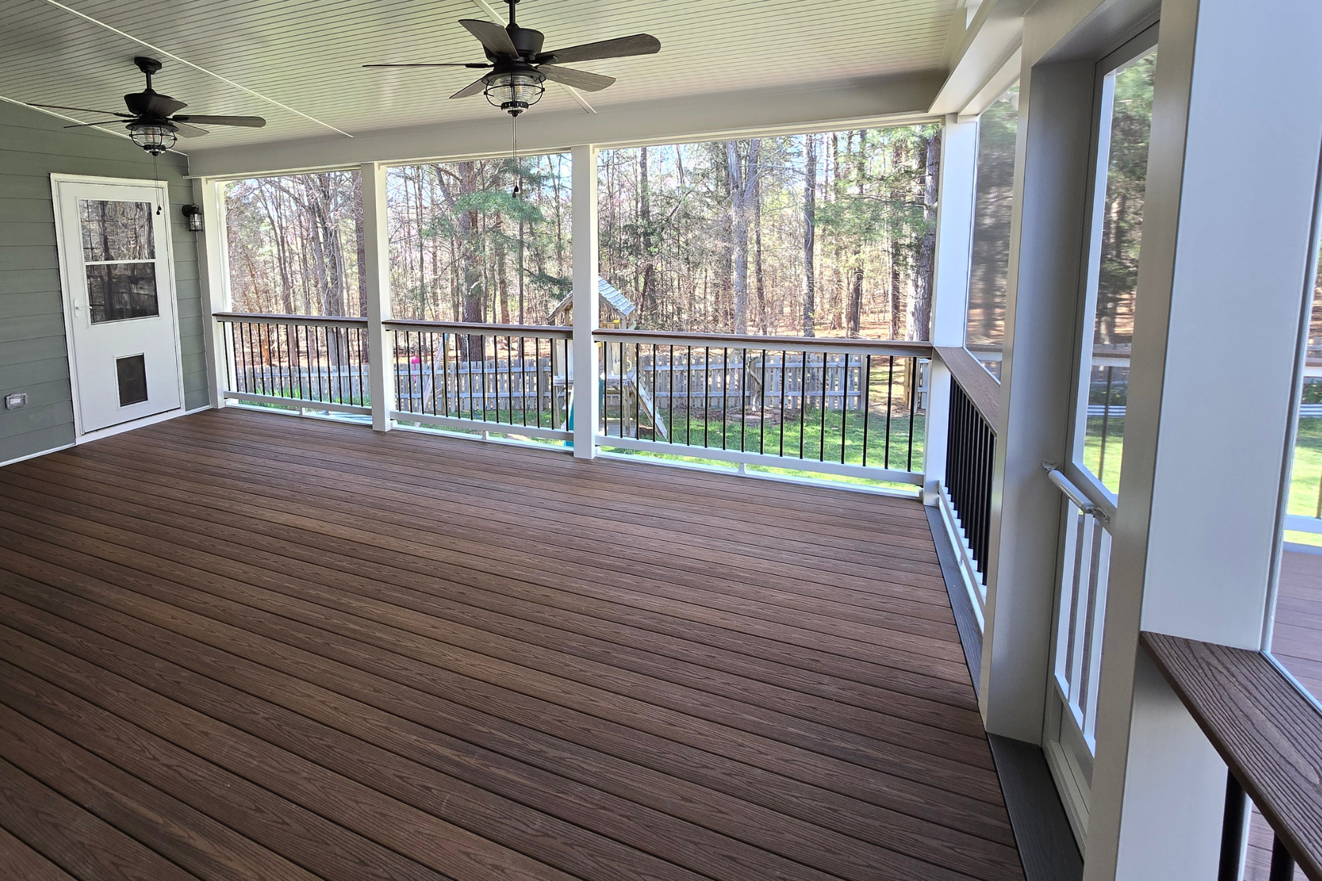 An empty covered wooden deck with a railing overlooking a wooded area, featuring ceiling fans and a door on the left.