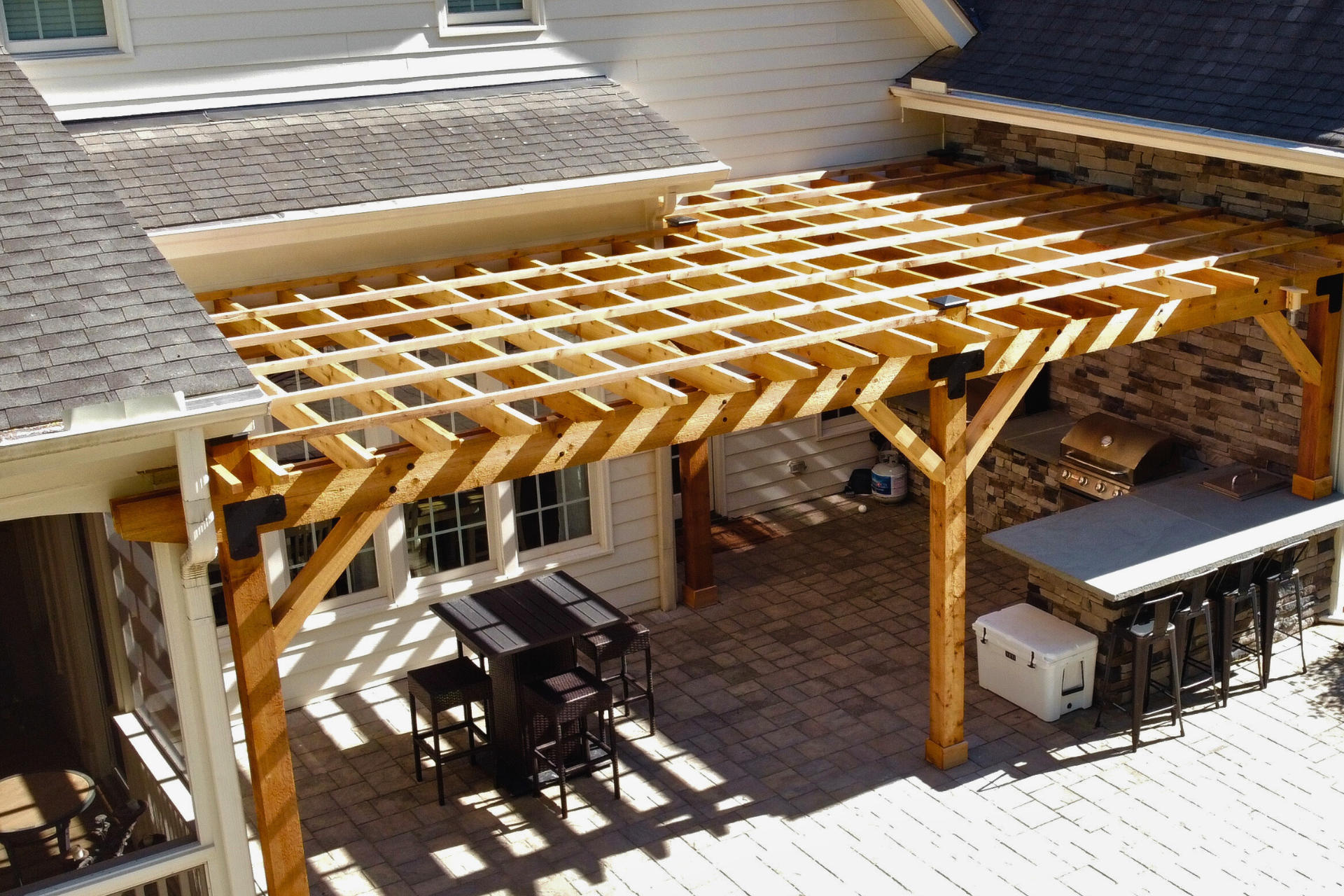A wood pergola covers a backyard patio with a dining set, an outdoor kitchen with a stone counter, and a white cooler.