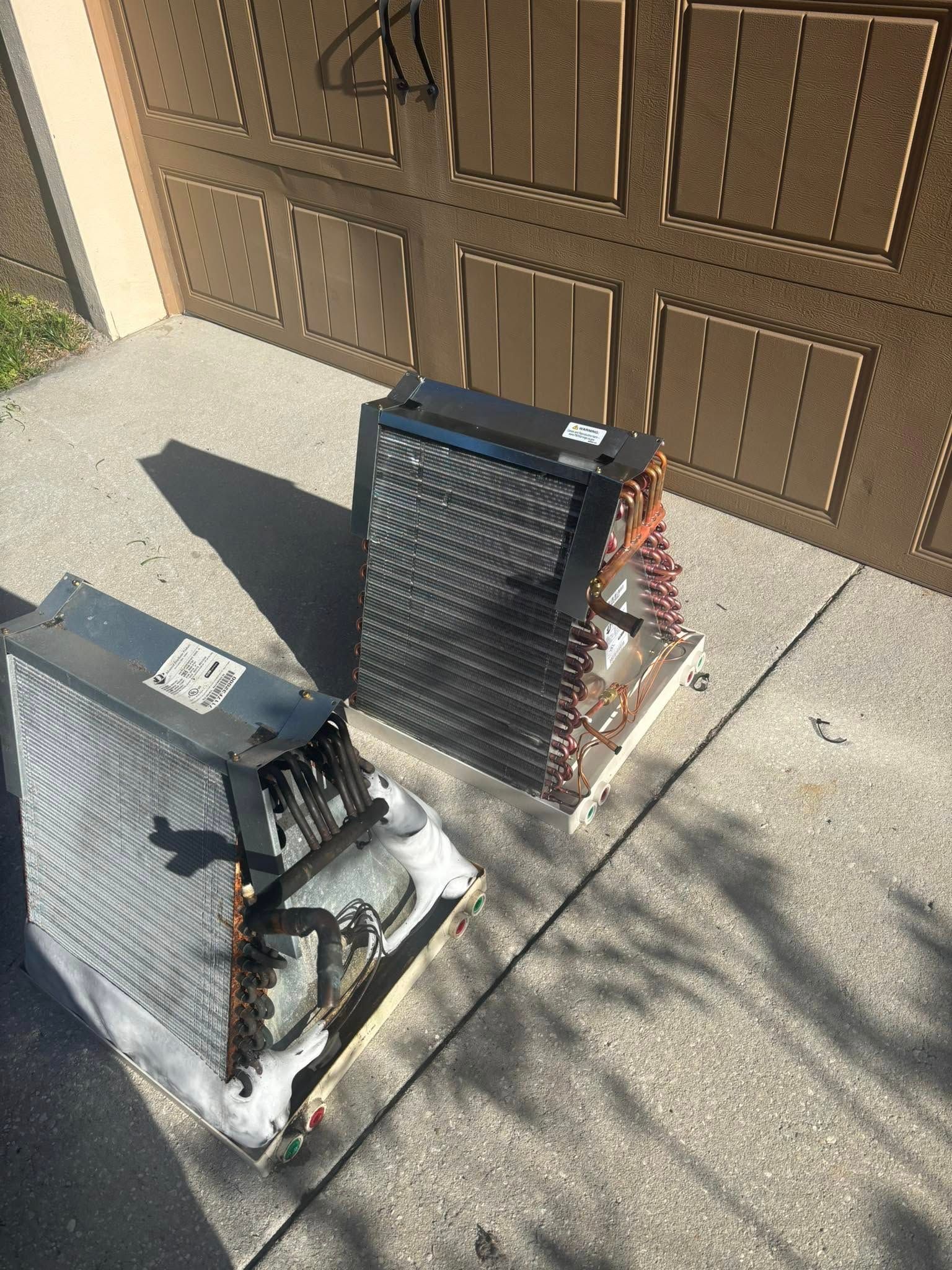 Two air conditioners are sitting on the sidewalk in front of a garage door.