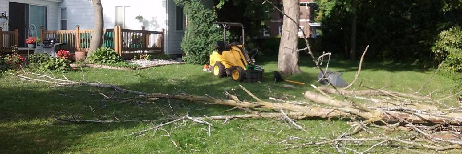 A lawn with a small yellow tractor, cut branches, and a house with a deck.