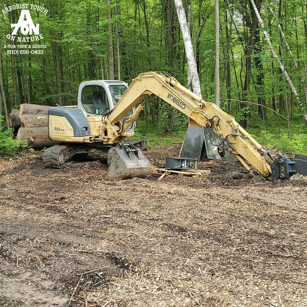 Yellow excavator in a wooded area, clearing wood chips.