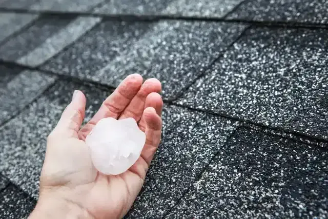 A person is holding a piece of hail in their hand in front of a roof.