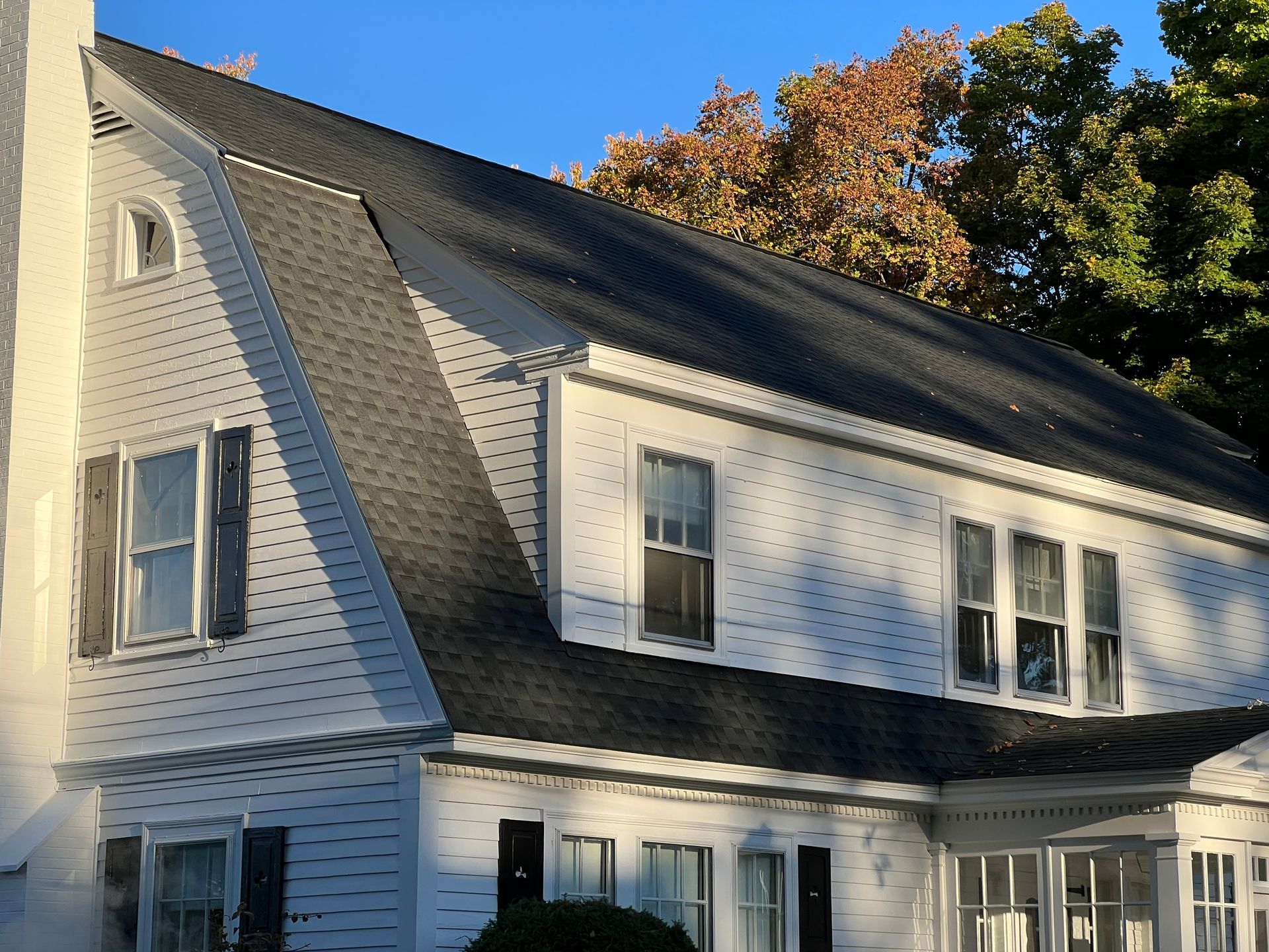 A white house with black shutters and a steep, dark-shingled gambrel roof, set against a backdrop of green trees.