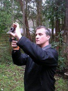 Man outdoors holding camera, aiming upwards, wearing a black jacket, among trees.