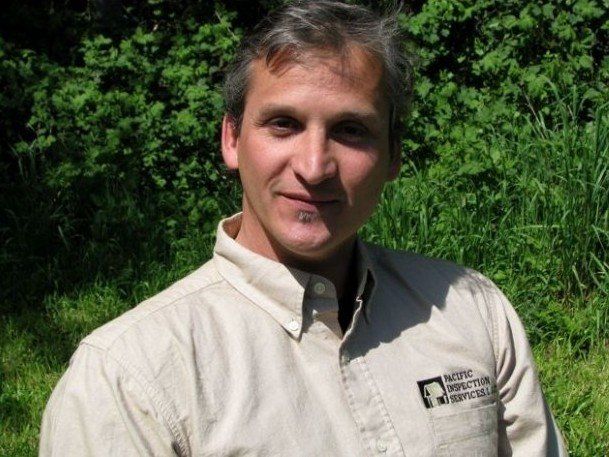 Man in a beige button-down shirt smiles, standing outdoors near green foliage.