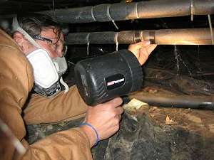 Person wearing a respirator examines pipes with a flashlight in a crawl space.